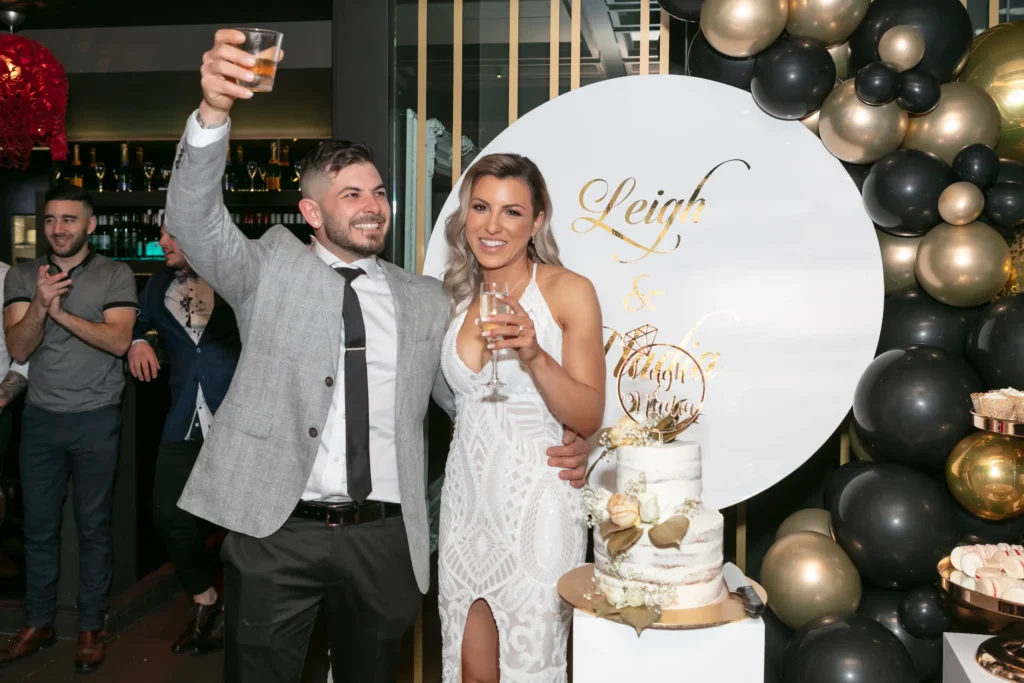 svg%3E A joyful engaged couple celebrates at their engagement party, standing front and center. The bride-to-be glows in a white halter-neck dress with lace details and a thigh-high slit, holding a champagne glass, while the groom-to-be raises his glass in a toast wearing a grey suit jacket and tie. Behind them, a large white sign with elegant gold script displays their names, and a multi-tiered white cake with floral accents sits nearby. Black and gold balloons complete the festive and elegant atmosphere, capturing the happiness and celebratory spirit of their special moment.