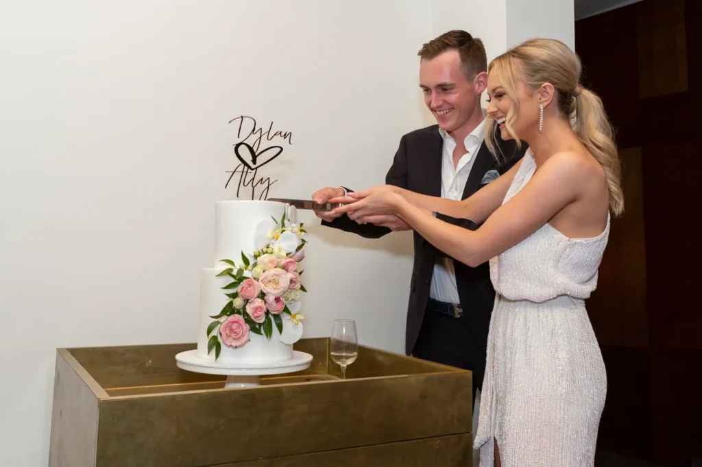 The engaged couple, Dylan and Ally, beam as they cut their white tiered engagement cake at a Melbourne engagement party. The cake is adorned with fresh pink and white flowers and green foliage, placed on a golden stand with a glass of champagne nearby. Their joyful expressions and elegant surroundings are beautifully captured in professional photography, highlighting the intimate, celebratory atmosphere of this special moment.