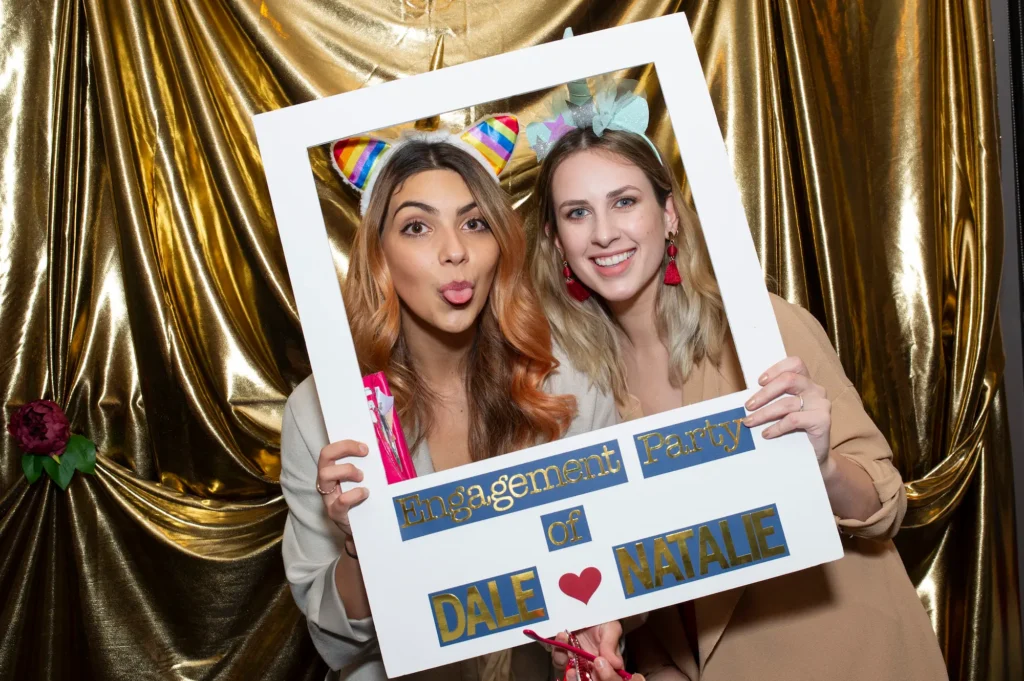 Two friends strike a playful pose in a custom photo booth frame at a Melbourne engagement party. One wears a rainbow-striped cat ear headband, the other a whimsical unicorn-inspired headband with tassel earrings. The frame reads "Engagement Party of DALE , NATALIE" in gold letters. A shimmering gold backdrop and a small floral decoration create a festive and glamorous setting. Professional photography captures the joy, friendship, and celebratory spirit of this memorable engagement party moment.