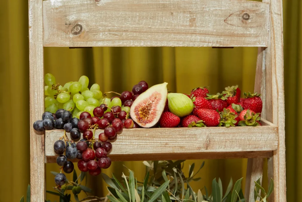 A beautifully arranged grazing table at a Melbourne engagement party features a rustic wooden tiered stand filled with fresh fruit. The top tier displays cascading green and red grapes, vibrant strawberries, and fresh figs, one sliced to reveal its pink interior. The lower tier is adorned with olive branches and green olives, adding a Mediterranean touch. Professional photography captures the colorful, fresh, and elegant presentation, highlighting the thoughtful and visually appealing details of the engagement party