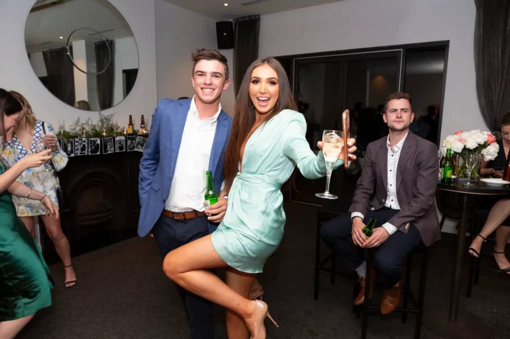Guests enjoy a lively moment at an engagement party. A woman in a pale mint green satin mini-dress poses playfully with a cocktail glass, while a man beside her smiles, dressed in a blue blazer and dark trousers holding a beer. Another seated guest observes, also holding a drink. The modern venue features a circular mirror, decorated mantel, and a table with bottles and floral arrangements. Professional photography captures the festive, stylish, and social atmosphere of friends celebrating together.