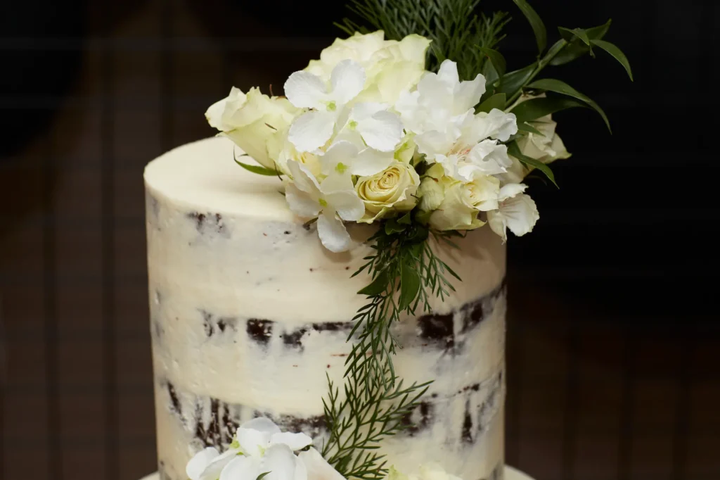 A close-up of a rustic-chic "naked cake" at engagement party, featuring multiple layers of dark cake with creamy white frosting between each layer. The unfrosted sides reveal the cake's natural texture, while the top is adorned with fresh white roses, smaller white blossoms, and cascading greenery. Professional photography captures the elegant, organic, and romantic presentation, highlighting the sophisticated simplicity of this intimate engagement celebration cake.
