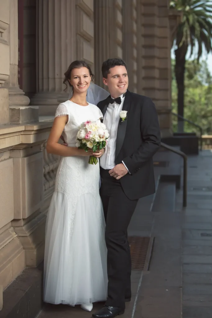 A newly married couple stands arm-in-arm in front of the grand Old Treasury Building in Melbourne, surrounded by classical stone columns and elegant architecture. The groom, dressed in a black tuxedo with a white boutonniere, looks lovingly toward his bride, who holds a bouquet of soft pink and white flowers. Her lace wedding gown glows in the natural sunlight, adding grace to the scene. The bright, clear day enhances the warmth and joy between them, creating a timeless and elegant wedding portrait that beautifully captures the romance and sophistication of a wedding in Melbourne’s historic Old Treasury Building.