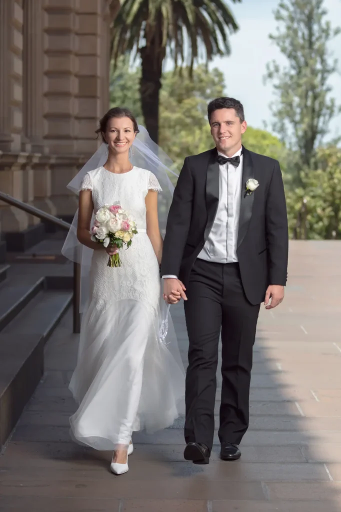 This joyful and formal wedding portrait captures a bride and groom walking hand-in-hand outdoors at the historic Old Treasury Building in Melbourne. The bride wears a flowing white lace gown with a long veil, holding a bouquet of blush, pink, and white roses, while the groom is in a classic black tuxedo with a bow tie. They smile confidently at the camera as sunlight highlights the architectural beauty of the building and lush greenery surrounding them. The image conveys the elegance, romance, and celebratory atmosphere of an Old Treasury Building wedding, creating a timeless and cinematic moment.