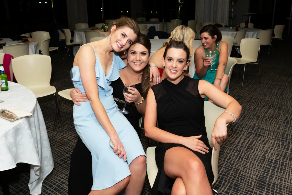 svg%3E Three young women enjoying Padraig Pearses GAC 50th Anniversary Awards Night at Zinc, Fed Square. One in a light blue dress leans forward smiling, another in a dark dress holds a glass of white wine, while a woman in black looks at the camera and another laughs in the background. Capturing candid, joyful moments among friends at a formal awards dinner, the warm lighting and elegantly set tables enhance the relaxed, celebratory atmosphere of this milestone Melbourne event, highlighting the camaraderie, laughter, and social connections of the evening.