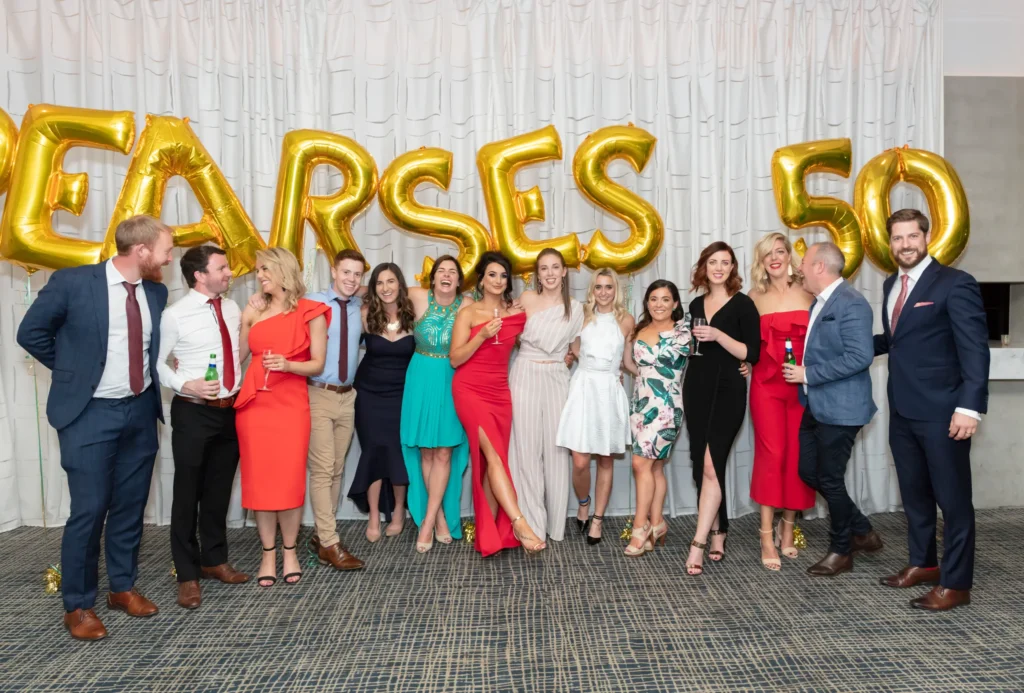 Group of 14 attendees celebrating Padraig Pearses GAC 50th Anniversary Awards Night at Zinc, Fed Square. Men in formal suits and women in elegant evening dresses in red, black, and teal smile, toast, and chat together. Behind them, large gold balloons spell 'PEARSES 50', highlighting the milestone celebration. The photograph captures the warmth, joy, and camaraderie of the evening, showcasing both posed and candid interactions as friends, colleagues, and community members come together to honor fifty years of achievement, spirit, and unity in Melbourne’s iconic waterfront venue.