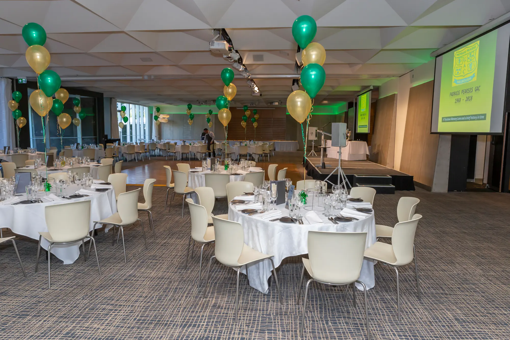 padraig pearses 50th anniversary zinc fed square room decor Empty event space at Zinc, Fed Square prepared for Padraig Pearses GAC 50th Anniversary Awards Night. Round tables with white tablecloths, plates, cutlery, glasses, and green centerpieces are neatly arranged, surrounded by modern white chairs. Bouquets of green and gold balloons float above tables, and a low stage with a microphone and projector screen displays 'PEARSES GAC 1964-2018'. Large windows allow natural light to fill the room, while ceiling light fixtures add elegance. This photograph captures the anticipation, refined decor, and meticulous attention to detail that set the stage for a formal, milestone celebration in Melbourne.