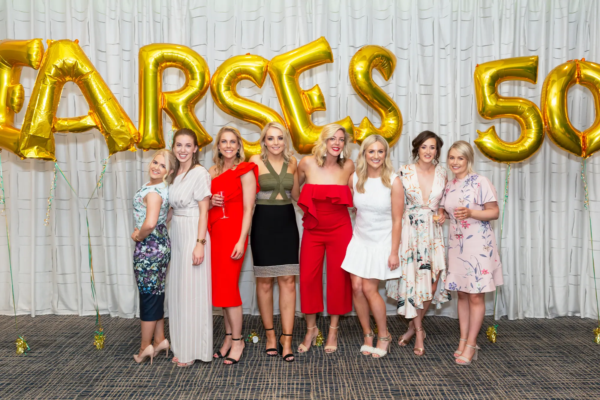 padraig pearses 50th anniversary zinc group photo women celebration Eight women stand side by side, posing for a celebratory group photo at the Pearses 50th Anniversary Awards Night. They wear a mix of colorful formal dresses and a jumpsuit, smiling and holding glasses in a playful, confident manner. Behind them, large gold balloons spell out "PEARSES 50," and gold streamers scatter across the dark patterned carpet. The backdrop of a simple white curtain contrasts with the lively expressions and stylish attire of the group, capturing a joyful moment of friendship, pride, and festive celebration during the milestone anniversary event.