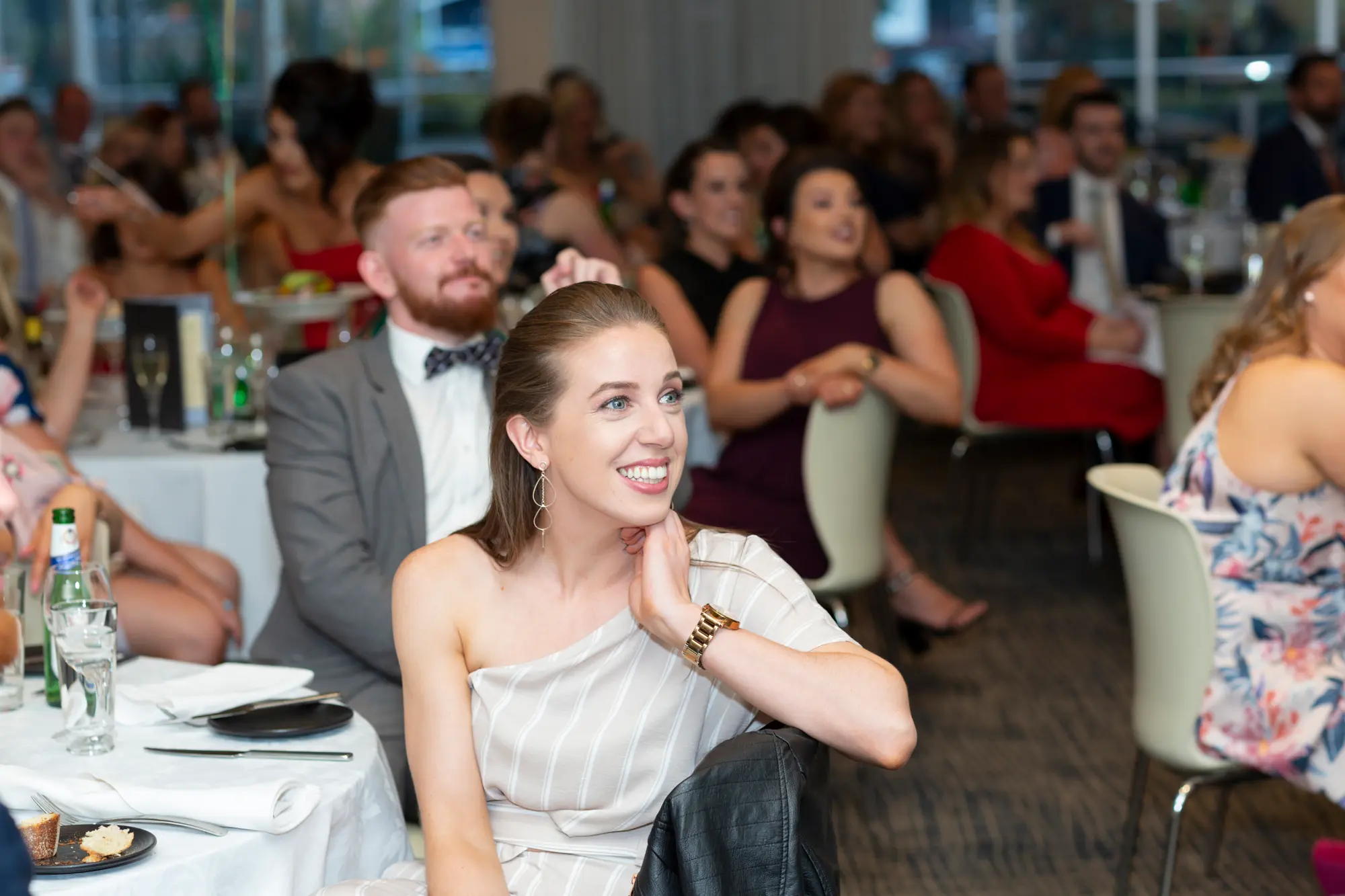 padraig pearses 50th anniversary zinc guests smiling during awards night This image captures a joyful and candid moment from the Pearses 50th Anniversary Awards Night. In the foreground, a woman with long hair wearing a striped, off-the-shoulder dress sits at a table, smiling brightly and resting her chin on her hand as she looks toward the stage. Her expression radiates happiness and engagement. Behind her, a man in a gray suit and bow tie is also smiling, watching the same direction. The background features elegantly set round tables with white tablecloths, glasses, and plates. The lighting is warm and soft, highlighting the cheerful and celebratory atmosphere of the evening.