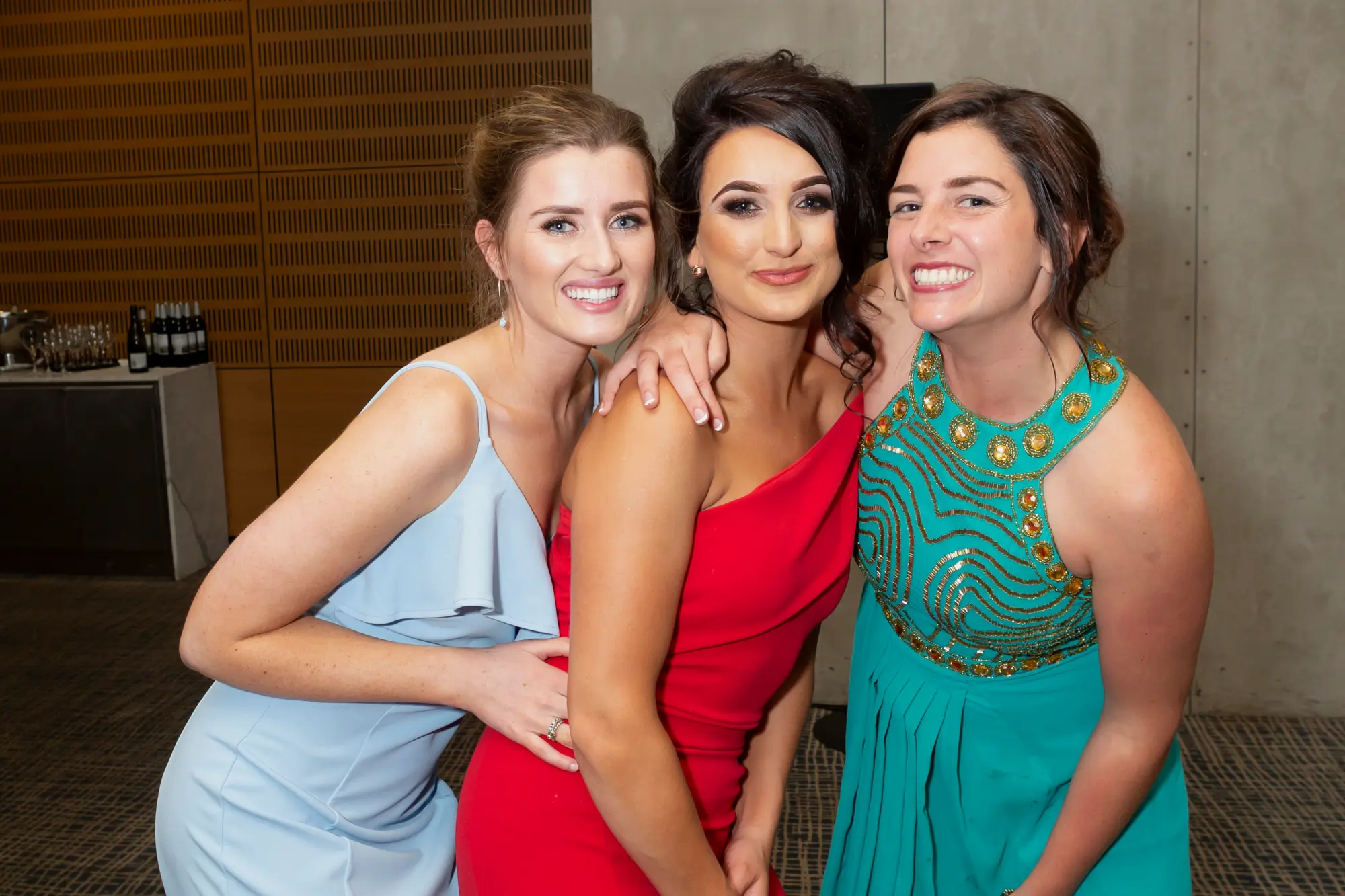 padraig pearses 50th anniversary zinc walking around three women celebration A candid, close-up moment of three women smiling and posing together during the Pearses 50th Anniversary Awards Night. The woman on the left wears a light blue ruffled dress, the center woman wears a red one-shoulder dress, and the woman on the right wears a teal halter dress with gold beading. They stand close together in a warm, social atmosphere, with a textured wooden wall and a table with drinks visible in the softly lit background, capturing the celebratory mood of the event.