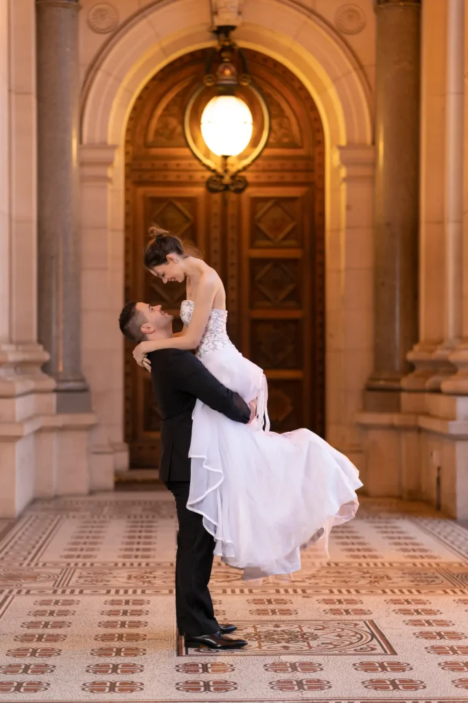 A joyful and dynamic indoor wedding portrait of a bride and groom at the Parliament House of Victoria in Melbourne. The groom lifts the bride off the ground as they share playful smiles, surrounded by grand architecture. A large, ornate wooden door with a circular glass window and fluted stone columns forms the dramatic backdrop, while the geometric tiled floor adds elegance. Warm lantern light casts a soft glow, highlighting the bride’s strapless beaded gown and flowing skirt and the groom’s classic black suit. The photograph captures the lively romance and celebratory energy of Melbourne bride and groom portraits during wedding photography.