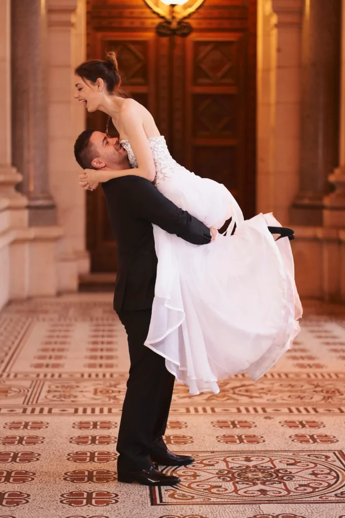 A joyful wedding moment captured inside the Parliament House of Victoria, where a groom lifts his bride into the air as they laugh together beneath grand architectural columns and a mosaic-tiled floor. The groom, dressed in a black suit, looks up at his bride with a bright smile, while her strapless white gown flows gracefully in motion. Warm natural light enhances the rich tones of the historic interior, highlighting the ornate wooden doors and elegant details. This candid and energetic scene reflects the happiness and romance of a wedding in Melbourne, beautifully showcasing authentic wedding photography at the Parliament House of Victoria.