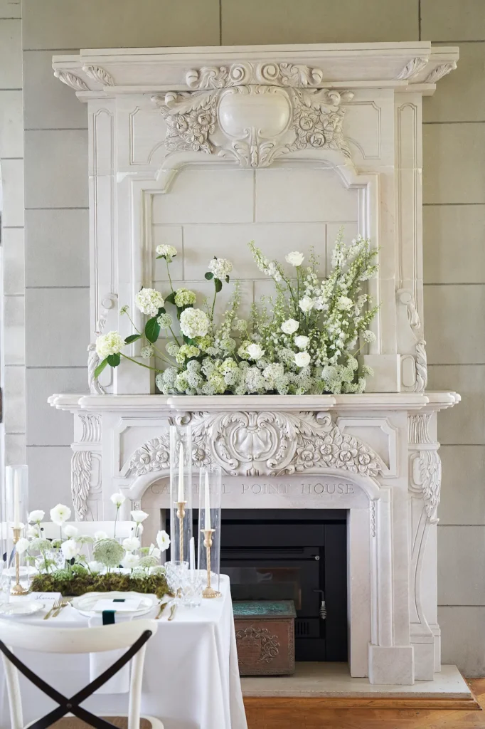 A grand, ornate fireplace mantel decorated with lush white and light green flowers serves as the focal point of a reception at Campbell Point House. The intricate carvings of the mantel are highlighted by soft natural light, while a portion of a nearby reception table is visible in the foreground, adorned with matching floral arrangements, tall glass candle holders, and gold-accented place settings. White chairs with X-shaped backs surround the table, contributing to the elegant and serene atmosphere. The scene captures a sophisticated, refined ambiance, emphasizing the beauty and attention to detail in this formal wedding reception setting.