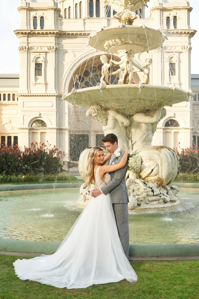 A sun-drenched wedding portrait of a bride and groom in front of the Royal Exhibition Building in Melbourne, captured during the golden hour. The couple stands close together on a vibrant green lawn, embraced under the warm, soft sunlight. A grand classical fountain with cherub sculptures flows behind them, and the historic building with arched windows and intricate details forms the majestic backdrop. The bride wears a sleeveless lace bodice gown with a flowing chiffon skirt, while the groom dons a light gray suit with a boutonnière. The photograph highlights the romantic intimacy of Melbourne bride and groom portraits during wedding photography.