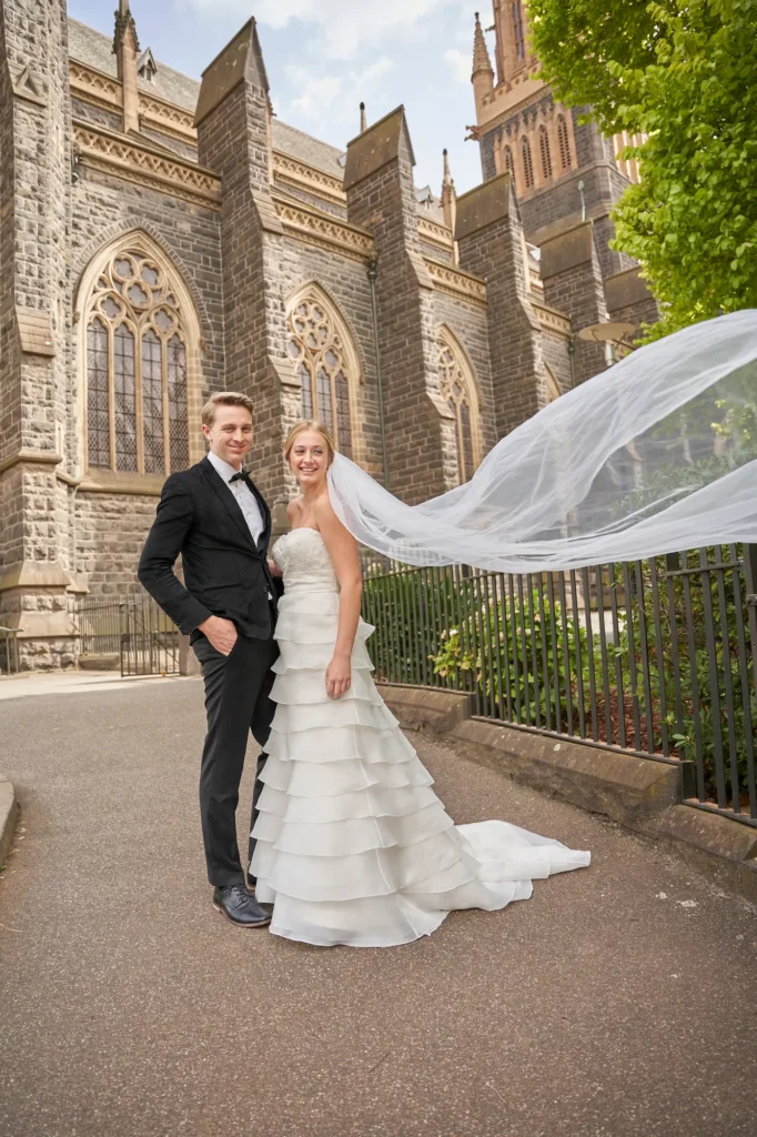 A newly married couple standing outside the grand St Patrick’s Cathedral in Melbourne for a timeless wedding portrait. The groom, dressed in a classic black suit and bow tie, stands confidently beside the bride, who wears a strapless white gown with a flowing veil caught by the wind. Behind them, the gothic-style stone facade of St Patrick’s Cathedral rises majestically, framed by lush greenery and soft daylight. The scene beautifully captures the elegance, romance, and architectural grandeur that define St Patrick’s Cathedral wedding photography in Melbourne.