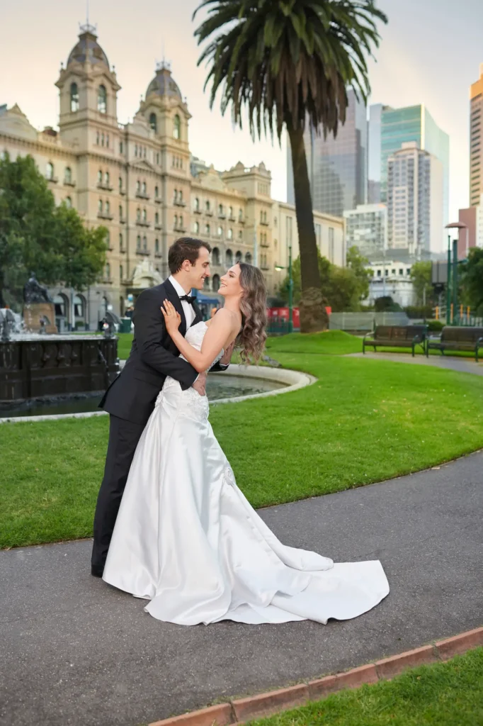 A bride and groom share an intimate embrace in Golden Reserve, with the elegant facade of The Hotel Windsor Melbourne rising behind them. The groom, dressed in a black tuxedo, holds his bride close as she leans gracefully into him, her strapless white gown glowing in the soft, golden afternoon light. Around them, the lush green lawns, a gentle fountain, and a tall palm tree frame the romantic moment, while the modern Melbourne skyline glimmers in the distance. This wedding Melbourne portrait beautifully captures the warmth, sophistication, and timeless love between the couple in an iconic city setting.