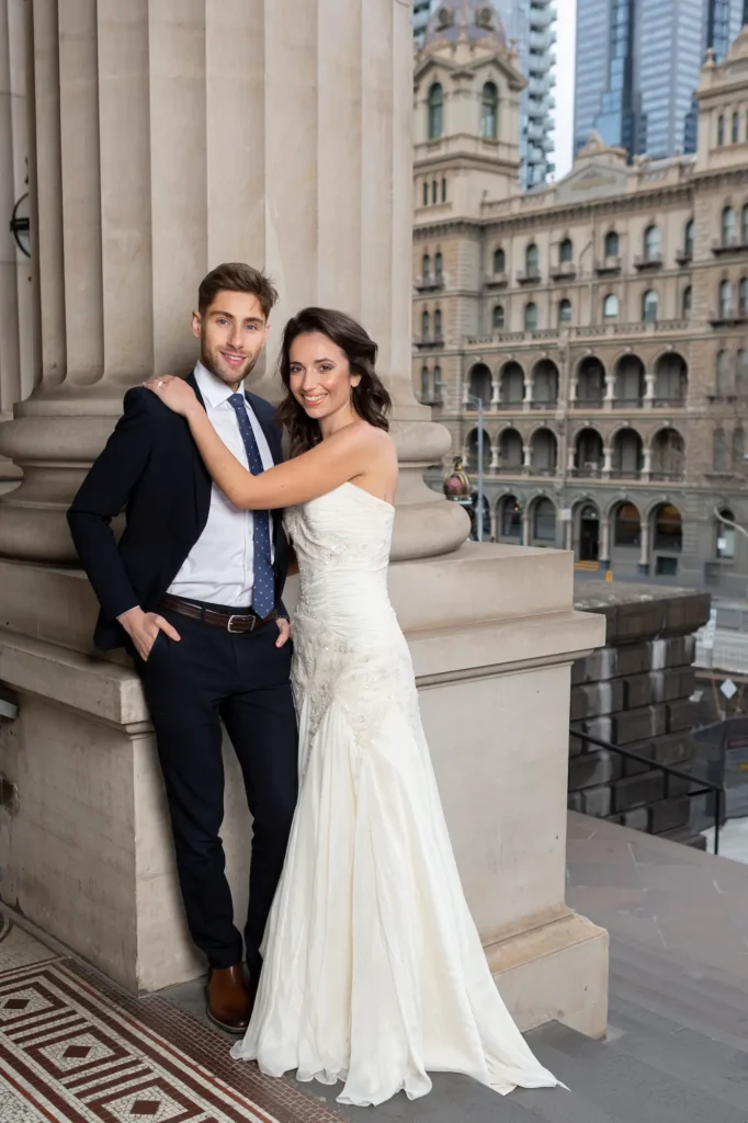A newly married couple posing for a classic wedding portrait at the Parliament House of Victoria in Melbourne. The groom, dressed in a dark suit and blue tie, leans casually against a grand stone column, while the bride, in a strapless white gown, stands beside him with a radiant smile. The background showcases the Parliament House’s elegant neoclassical architecture, with tall fluted columns and arched windows, along with a glimpse of The Hotel Windsor in the distance. The bright daylight enhances the refined and celebratory mood, perfectly capturing the timeless elegance of Parliament House of Victoria wedding photography in Melbourne.