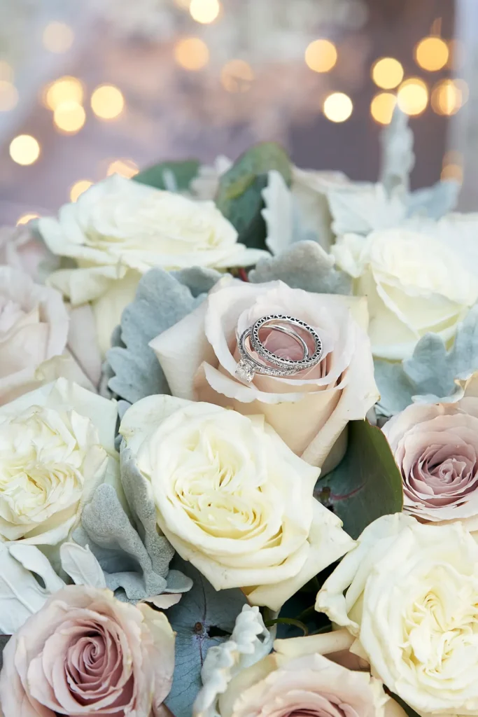 A romantic close-up photograph capturing the bride’s wedding rings nestled gently within the petals of a soft blush-pink rose. The engagement ring, with a brilliant center diamond, rests atop a diamond-studded wedding band, both shimmering in the delicate light. Surrounding them is a bouquet of roses in creamy white, pale pink, and muted champagne tones, accented with touches of green foliage. The softly blurred background with warm bokeh adds an intimate, dreamy atmosphere. This detailed shot from the bride getting ready captures the elegance, love, and anticipation of a Melbourne wedding morning.
