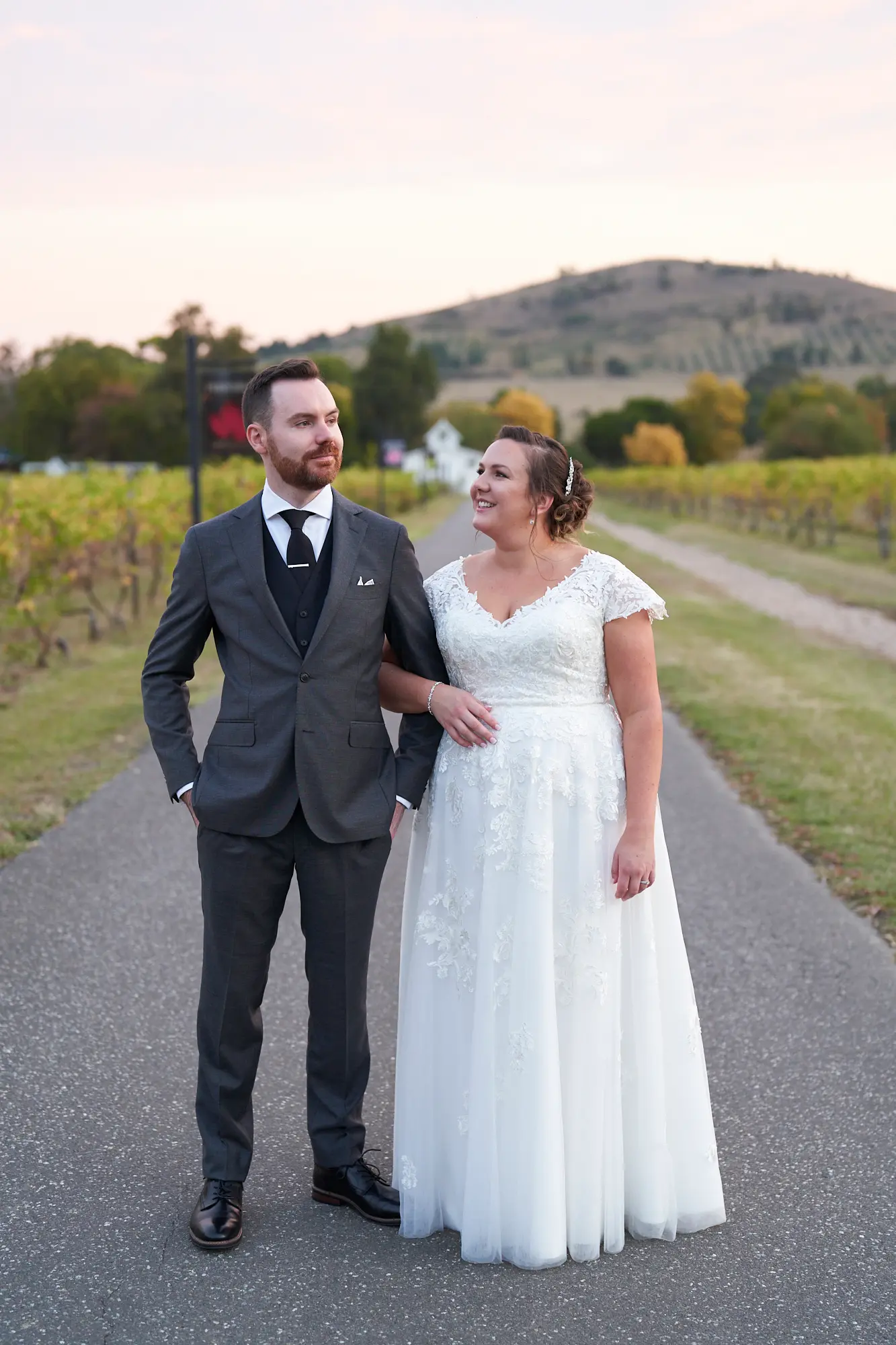 erin jesse arm in arm driveway fergusson winery yarra valley wedding This wedding photo captures the bride and groom walking arm-in-arm along Fergusson Winery’s scenic driveway in the Yarra Valley. They gaze at each other with wide, joyful smiles, radiating love and excitement. Rows of autumn-toned grapevines frame the paved path, while a white farmhouse with a red roof and rolling hills create a picturesque rural backdrop. A rustic wooden winery sign adds charm, and scattered trees enhance the natural landscape. The couple’s elegant attire and relaxed pose blend seamlessly with the vineyard’s rustic elegance, creating a warm, intimate, and timeless portrait of their special day.