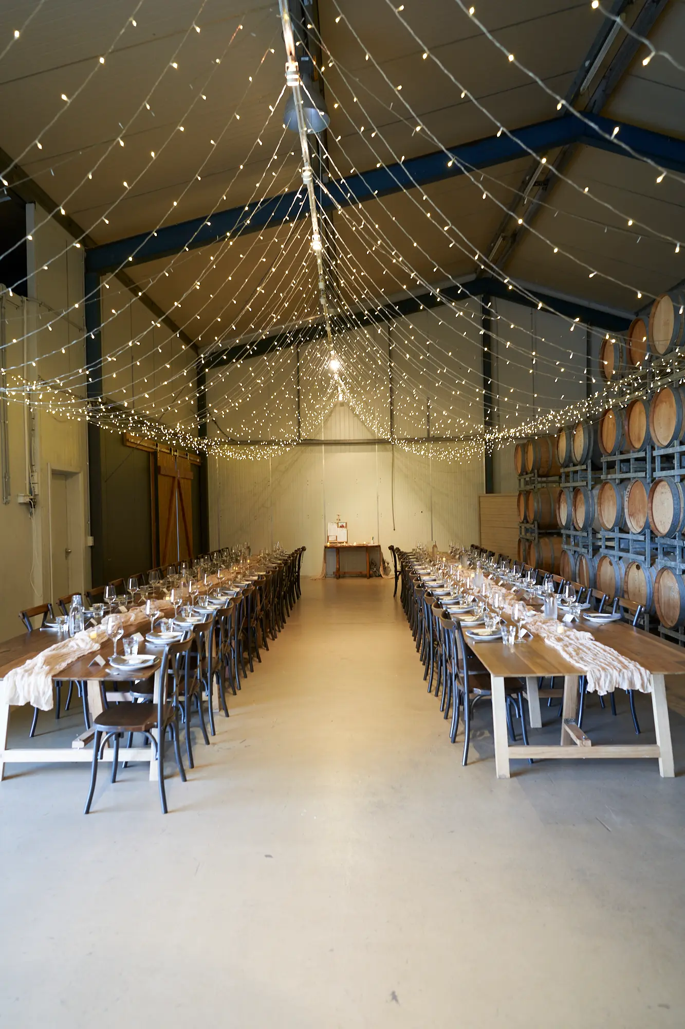 erin jesse barrel room reception fergusson winery This photo captures the barrel room at Fergusson Winery elegantly set for a wedding reception, blending rustic charm with refined detail. Long wooden tables are perfectly arranged with white plates, glassware, and light runners, ready for guests. Stacked wooden wine barrels line the walls, and twinkling string lights hang from the industrial ceiling, casting a warm, inviting glow. The polished concrete floor and open space balance the cozy ambiance. This serene, paused moment evokes anticipation and excitement for the celebration, highlighting the winery’s unique combination of rustic authenticity and elegant wedding styling in a welcoming atmosphere.