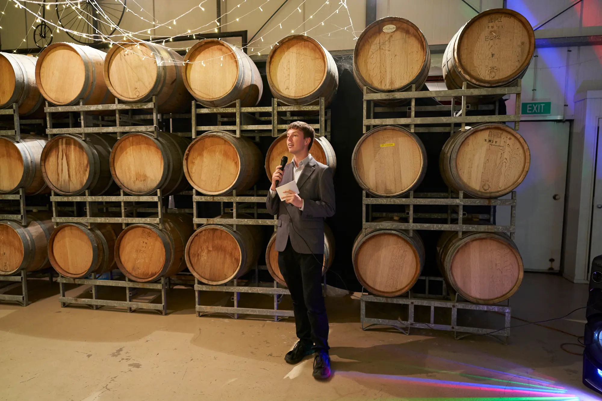 erin jesse barrel room speech fergusson winery yarra valley wedding The bride’s brother gives a warm and engaging speech during the wedding reception in Fergusson Winery’s barrel room. Standing confidently in a dark gray suit, he holds a microphone and notes while smiling at guests. Stacked wooden wine barrels line the walls, and hundreds of twinkling string lights create a cozy, intimate, and celebratory ambiance, capturing the emotional and rustic elegance of the winery setting.