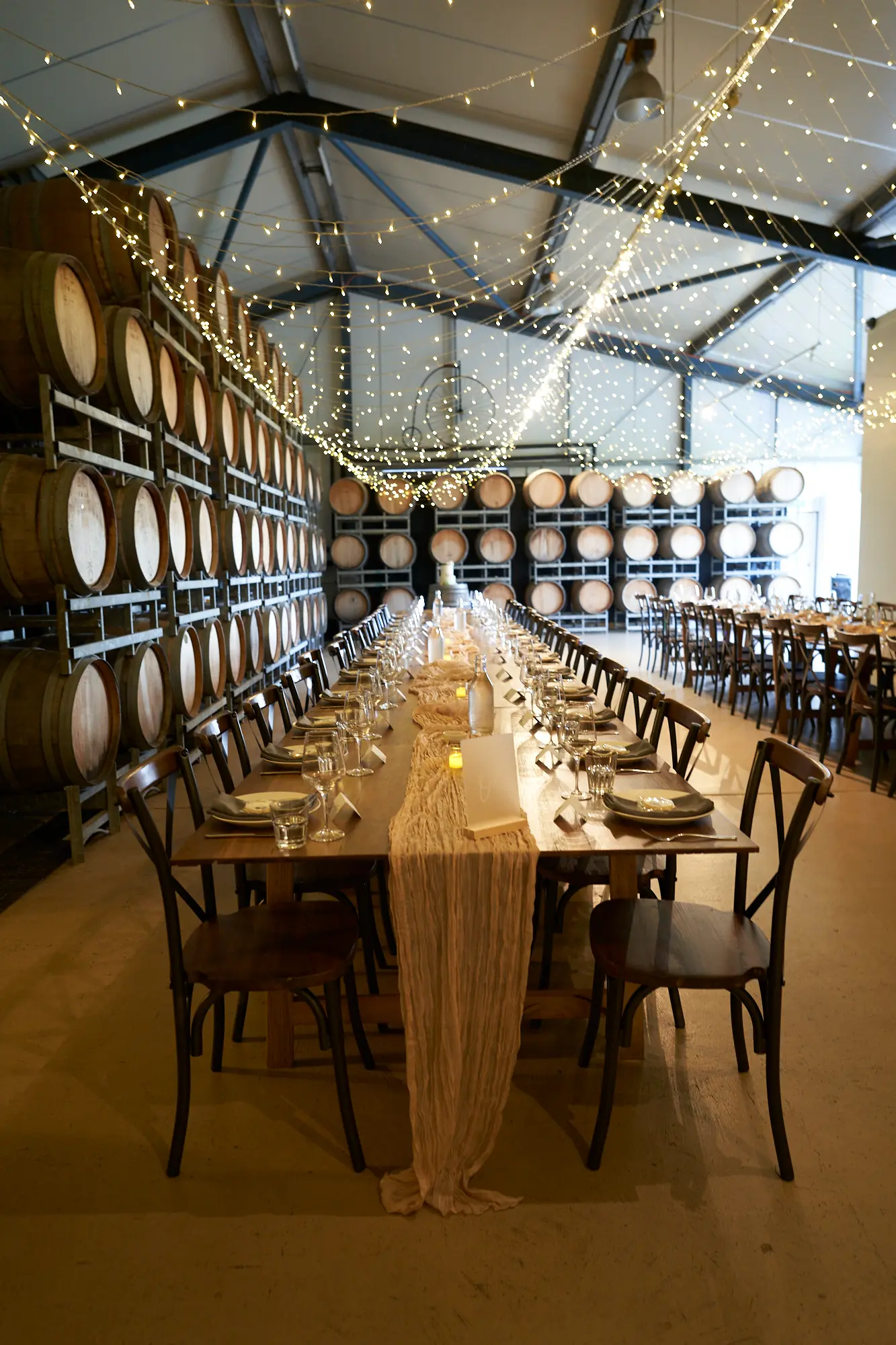 erin jesse barrel room wedding reception fergusson winery This photo showcases the rustic elegance of Fergusson Winery’s barrel room, fully set for a wedding reception. A long, dark wooden table stretches down the center, lined with cross-back chairs and adorned with white plates, glassware, and a light table runner. Stacked wooden wine barrels frame the sides, adding warmth and authenticity. Hundreds of twinkling string lights hang from the industrial ceiling, casting a cozy glow across the polished concrete floor. The room exudes anticipation and intimacy, blending the raw textures of the working winery with refined details, ready for guests to celebrate in style.