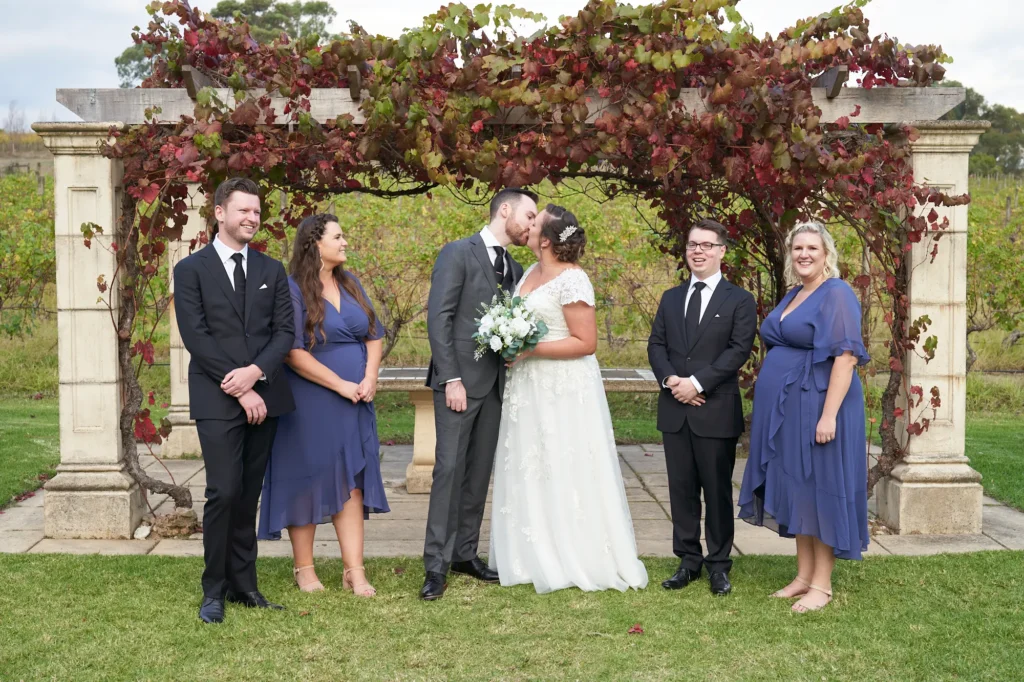erin jesse bridal party kiss under gazebo fergusson winery yarra valley wedding A joyful bridal party portrait at Fergusson Winery in the Yarra Valley, featuring the bride and groom sharing a tender kiss under a rustic sandstone gazebo draped in red and green autumn grapevines. Flanking them, two bridesmaids in dusty blue dresses and two groomsmen in dark suits smile warmly at the camera. Behind them, rows of vineyards and rolling hills stretch into the distance under a bright, partly cloudy sky. The scene captures intimate celebration, rustic elegance, and collective happiness of the wedding party on this picturesque outdoor wedding day.