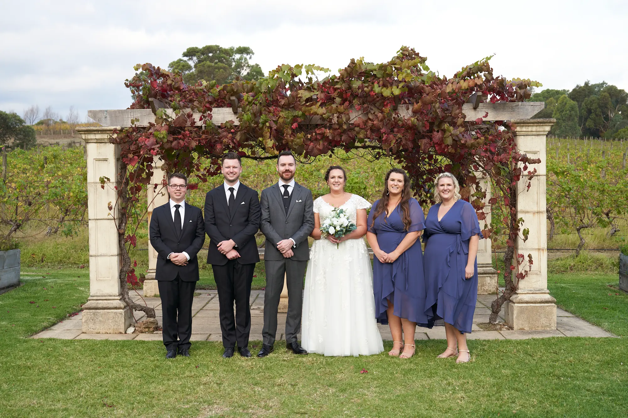 erin jesse bridal party sandstone gazebo fergusson winery yarra valley wedding The bride, groom, and four bridal party members pose for a joyful group portrait at Fergusson Winery’s Sandstone Gazebo in the Yarra Valley. The couple stands at the center, flanked by two bridesmaids in dusty blue dresses and two groomsmen in dark suits, all smiling warmly at the camera. Behind them, the rustic sandstone gazebo is draped in lush grapevines with autumn red and green leaves, with vineyards and rolling hills stretching into the distance, capturing a festive, intimate, and scenic wedding moment full of warmth, camaraderie, and rustic elegance.