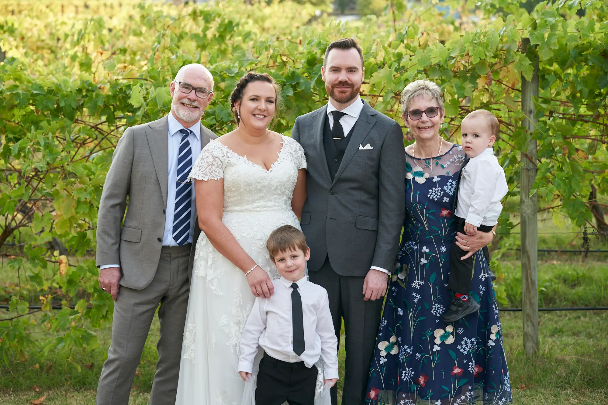 erin jesse bride groom groom family fergusson winery yarra valley wedding The bride and groom pose outdoors with the groom’s parents and their two young sons at Fergusson Winery, Yarra Valley. The couple stands in the back row, smiling warmly, flanked by the groom’s parents, while the older son sits on the ground and the toddler is held by the groom’s mother. Behind them, lush vineyard rows with green and yellow autumn leaves create a rustic, scenic backdrop. The photograph captures a joyful, three-generation family portrait, radiating warmth, celebration, and elegant rustic charm in a tranquil outdoor wedding setting.