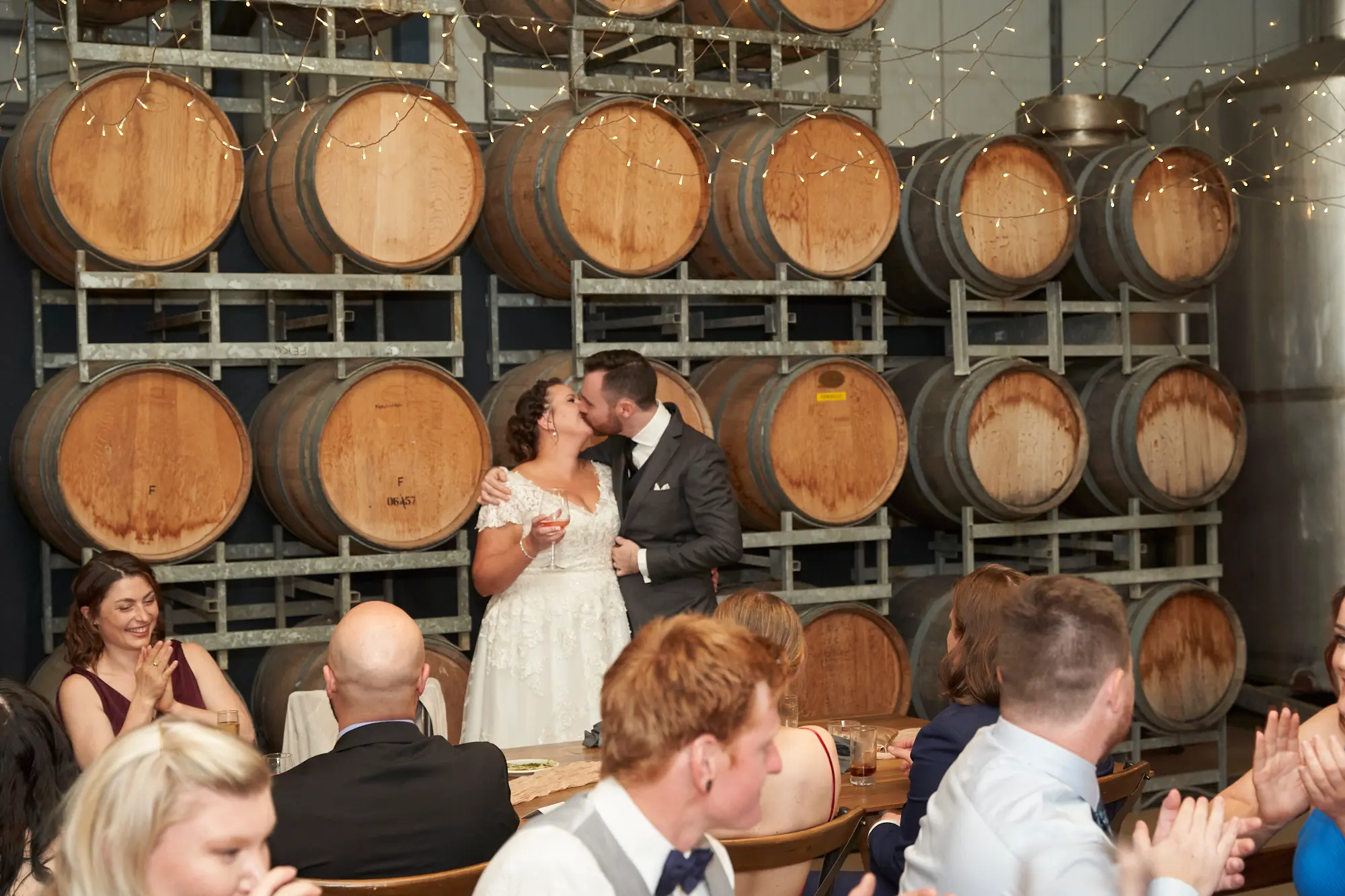 erin jesse bride groom kiss barrel room fergusson winery yarra valley wedding The bride and groom share a tender kiss in the middle of Fergusson Winery’s barrel room after a speech, with the groom’s arm around her waist and her hands on his cheeks. Guests at rustic wooden tables watch, smile, and clap in celebration. Stacked wine barrels and a stainless steel tank frame the warm, intimate space, while hundreds of twinkling string lights overhead cast a soft, cozy glow, creating a joyful and rustic-chic atmosphere that highlights the couple’s love and the personal, heartfelt moments of their winery wedding reception.