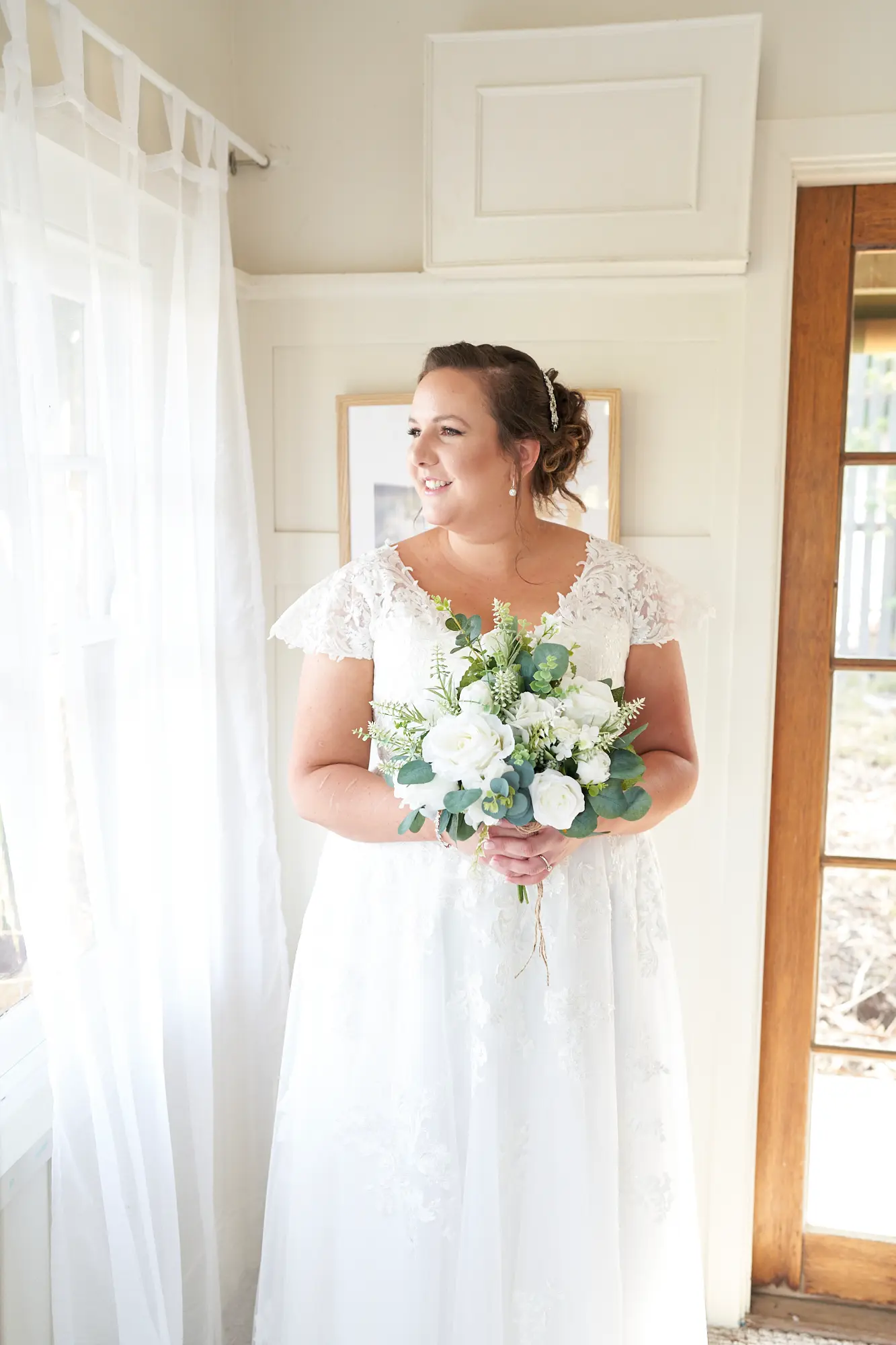 erin jesse bride standing guesthouse fergusson winery yarra valley Bride standing gracefully in a bright guesthouse at Fergusson Winery in the Yarra Valley, holding a white and green floral bouquet. She gazes off-camera with a warm, natural smile, bathed in soft natural light streaming through sheer curtains. The elegant lace gown, relaxed posture, and serene indoor setting create a calm, intimate, and joyful moment of anticipation before the wedding ceremony, highlighting the peaceful preparation, personal reflection, and timeless beauty of the bridal experience in the charming winery accommodation.