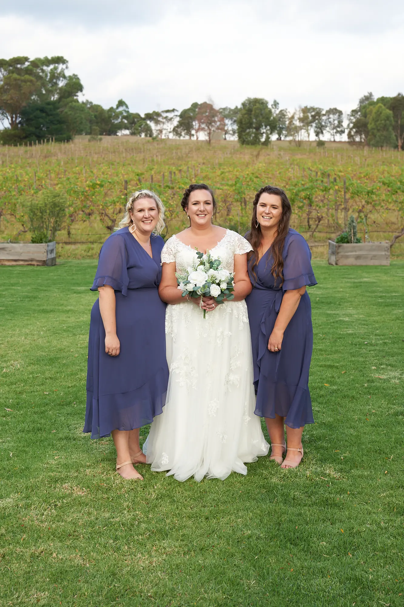 erin jesse bride with bridesmaids fergusson winery yarra valley wedding A joyful bridal party portrait at Fergusson Winery in the Yarra Valley featuring the bride standing between her two bridesmaids on a lush green lawn. The bride wears a white lace dress and holds a white and green bouquet, while the bridesmaids wear matching navy blue flowing dresses. Behind them, rows of grapevines and rolling hills provide a scenic vineyard backdrop. The bright daylight, warm smiles, and relaxed poses capture the happiness, camaraderie, and rustic elegance of this special wedding day.