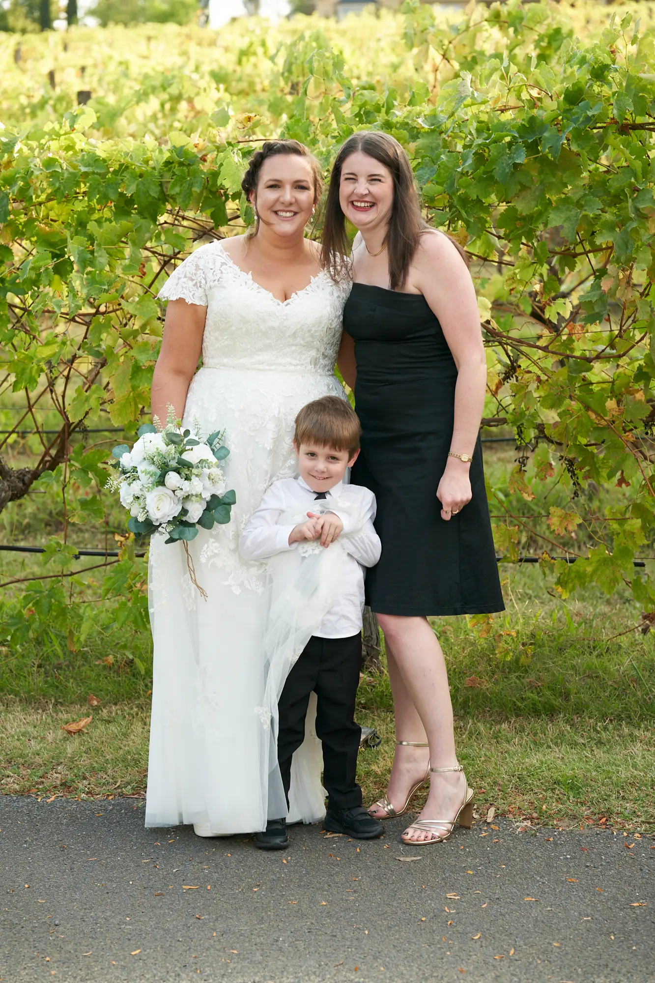 erin jesse bride with friends fergusson winery yarra valley wedding The bride stands outdoors at Fergusson Winery with a young boy and a female friend, posing for a warm, joyful group portrait. She holds a white and green floral bouquet, smiling brightly at the camera, while her companions flank her, sharing happy expressions. Behind them, rows of lush grapevines in green and yellow autumn hues stretch into the distance, set against the scenic Yarra Valley. The bright natural light and vineyard backdrop create an intimate, elegant, and celebratory atmosphere, capturing a personal moment of happiness with close friends during the wedding day.