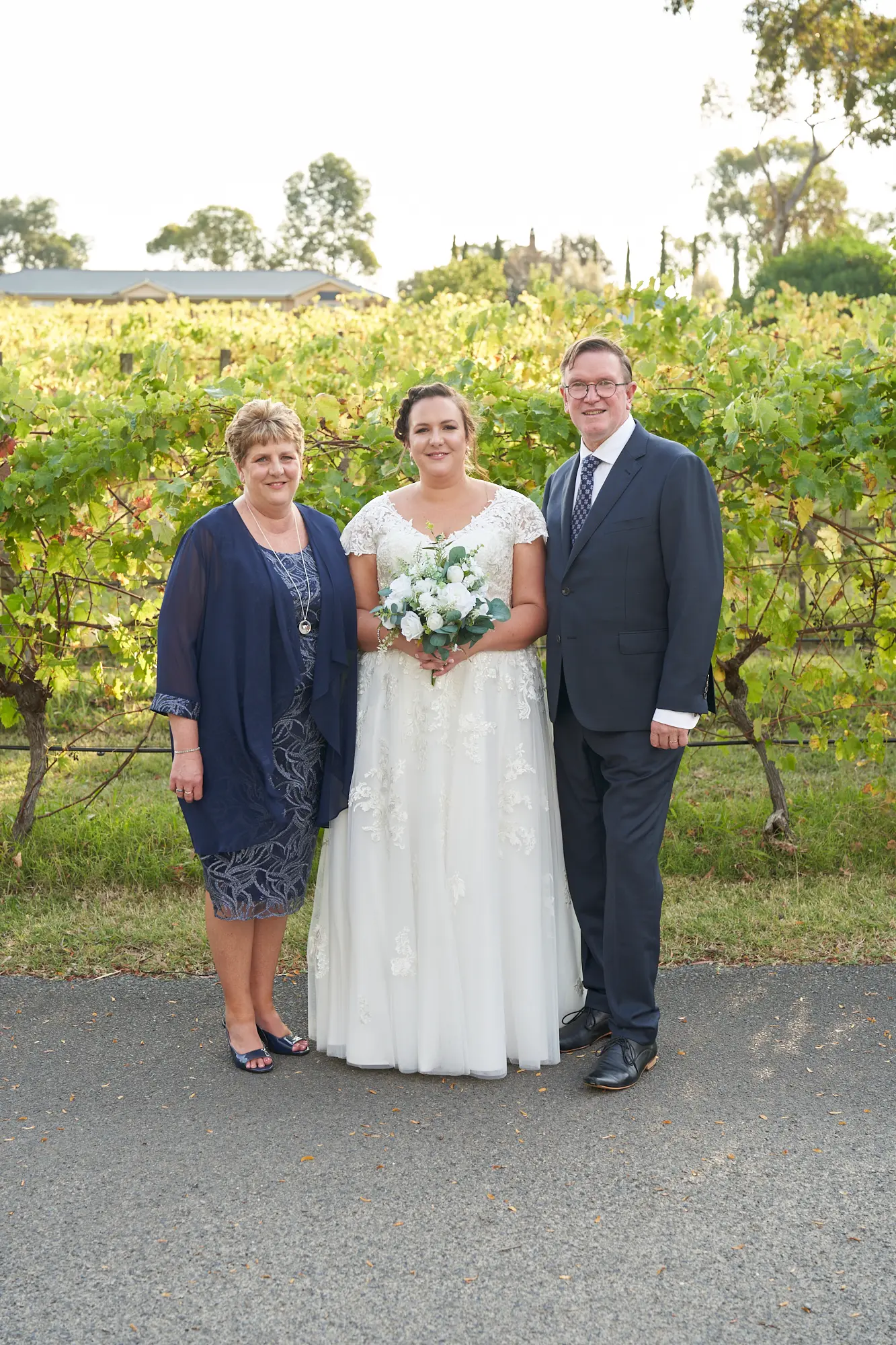 erin jesse bride with parents fergusson winery yarra valley wedding The bride stands outdoors at Fergusson Winery with her parents, posing for a warm and joyful family portrait. She wears a white lace wedding dress and holds a white and green floral bouquet, smiling brightly at the camera. Her mother, in a navy blue dress, and her father, in a dark gray suit, flank her on either side. Behind them, rows of lush grapevines in green and yellow autumn hues create a rustic vineyard backdrop, with distant winery buildings and trees completing the scenic Yarra Valley setting. The image conveys intimacy, elegance, and celebratory family joy.