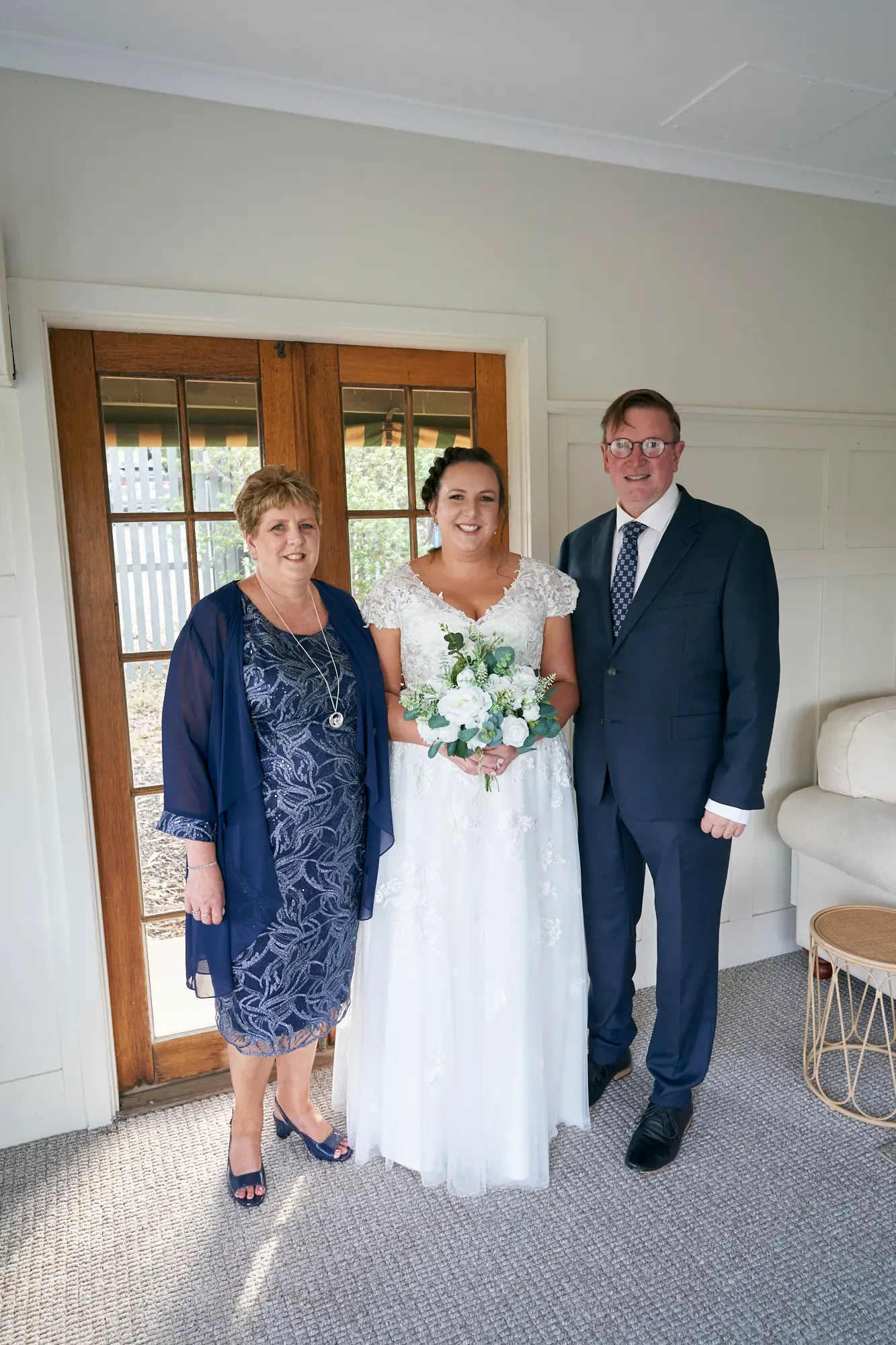 erin jesse bride with parents guesthouse fergusson winery yarra valley Bride posing with her parents for a joyful family portrait in a bright guesthouse room at Fergusson Winery in the Yarra Valley. The bride stands between her mother in a navy blue dress and her father in a dark suit, holding a white and green floral bouquet and smiling warmly at the camera. Natural light illuminates the light-colored walls, white wainscoting, and wooden-framed glass doors, creating an intimate, celebratory, and proud moment that captures the close family bond and happiness on the wedding day.