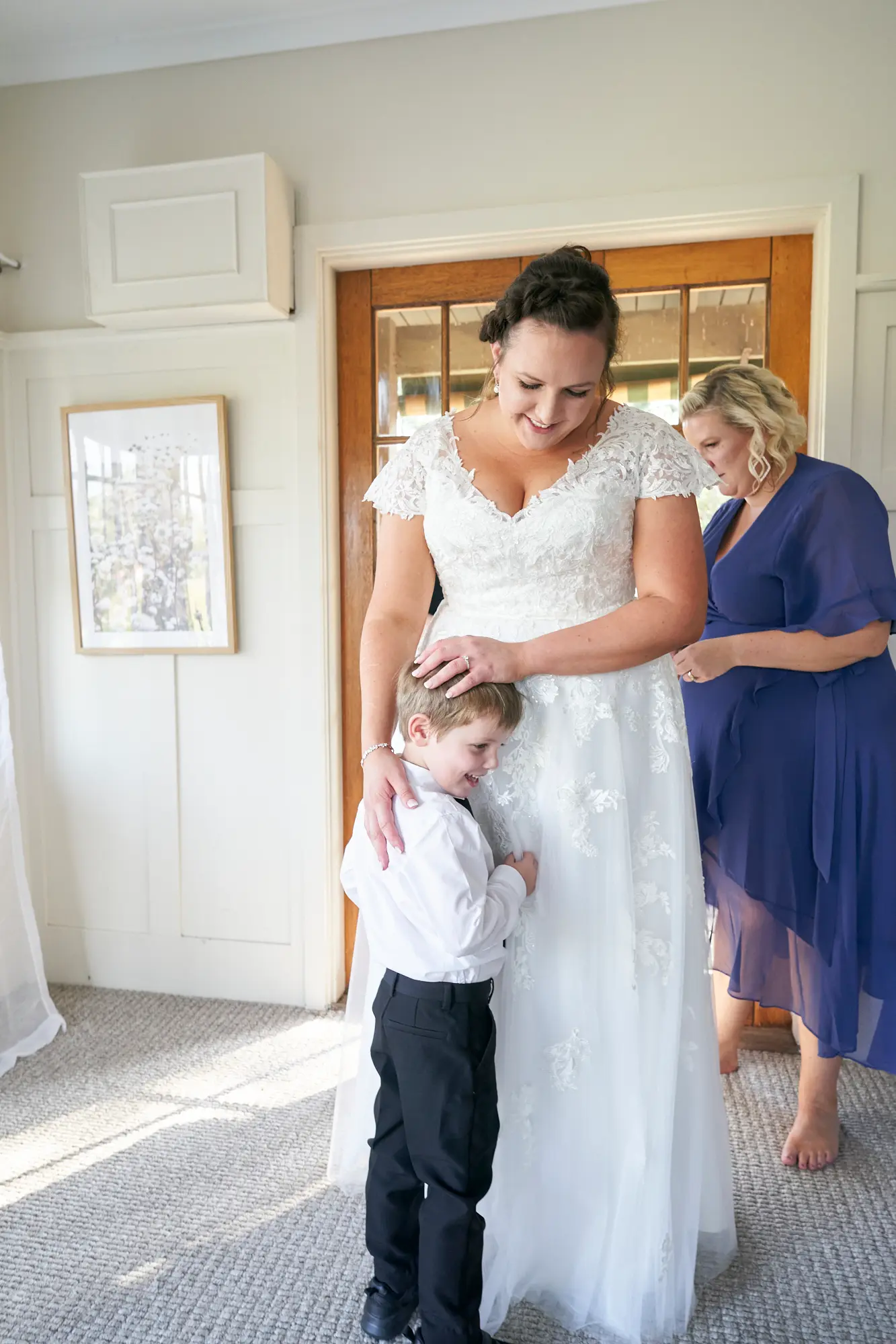 erin jesse bride with young boy guesthouse fergusson winery yarra valley Bride sharing a tender moment with a young boy in a bright guesthouse room at Fergusson Winery in the Yarra Valley. The boy rests his head against her white wedding dress and wraps his arms around her waist, while the bride looks down at him with warmth. Natural light fills the room with light-colored walls, white wainscoting, a carpeted floor, and wooden-framed glass doors, creating an intimate, serene, and heartfelt pre-ceremony moment that highlights the quiet joy and emotional connections of the wedding day.
