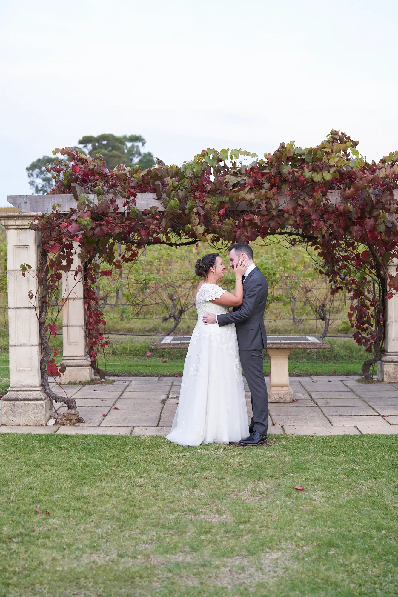 erin jesse close embrace sandstone gazebo fergusson winery yarra valley wedding This wedding photo captures a tender and intimate moment as the bride and groom stand closely, gazing into each other’s eyes under a vine-draped sandstone gazebo at Fergusson Winery in the Yarra Valley. Their embrace, with the groom’s arm around the bride’s waist and her hands on his cheeks, creates a personal, heartfelt connection. Framed by autumn-toned grapevines and lush vineyards, this serene portrait blends rustic elegance with natural beauty, highlighting their love and joy during a quiet, private moment in their outdoor wedding portrait session.