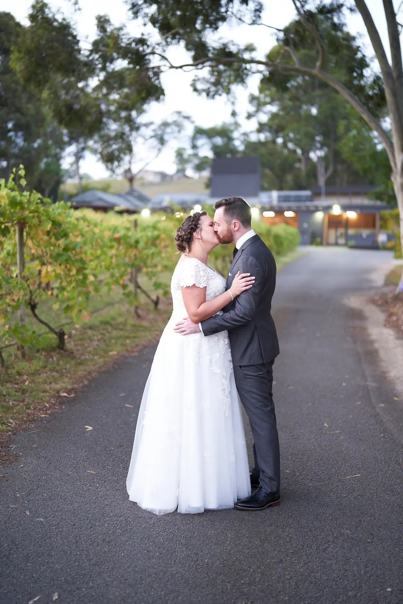 erin jesse couple kiss embrace paved path fergusson winery yarra valley wedding This wedding photo captures an intimate and romantic moment as the bride and groom share a gentle kiss on a paved driveway at Fergusson Winery in the Yarra Valley. The groom’s arm wraps around the bride’s waist while her hands rest tenderly on his cheeks, highlighting their deep connection. To the left, rows of vibrant vineyards stretch along the edge, while the winery’s elegant building with warm exterior lights stands in the background. Large trees frame the scene, adding depth and natural beauty. The image exudes romantic serenity, rustic elegance, and a private, heartfelt moment between the couple.