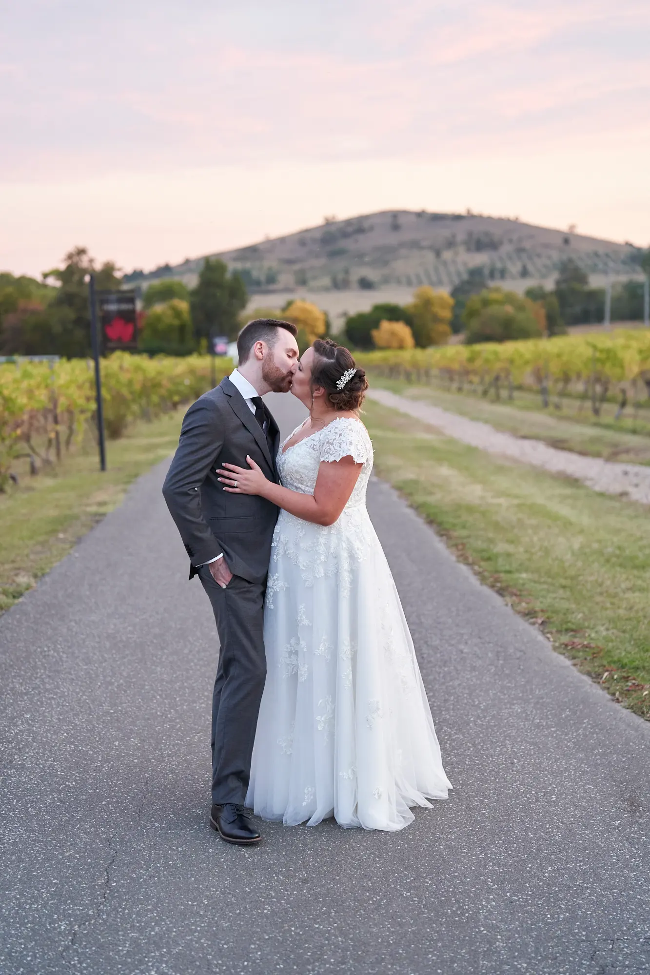 erin jesse driveway kiss fergusson winery yarra valley wedding This wedding photo captures the bride and groom sharing a gentle kiss in the middle of Fergusson Winery’s paved driveway in the Yarra Valley. They stand face-to-face in a close embrace, the groom’s hand in his pocket and his arm around her waist, while the bride’s hand rests on his chest. Rows of autumn-toned grapevines frame the path, with rolling hills and scattered trees creating a serene rural backdrop. The couple’s elegant attire and intimate pose convey deep love and joy, blending rustic vineyard charm with romantic, timeless elegance during their private portrait session.