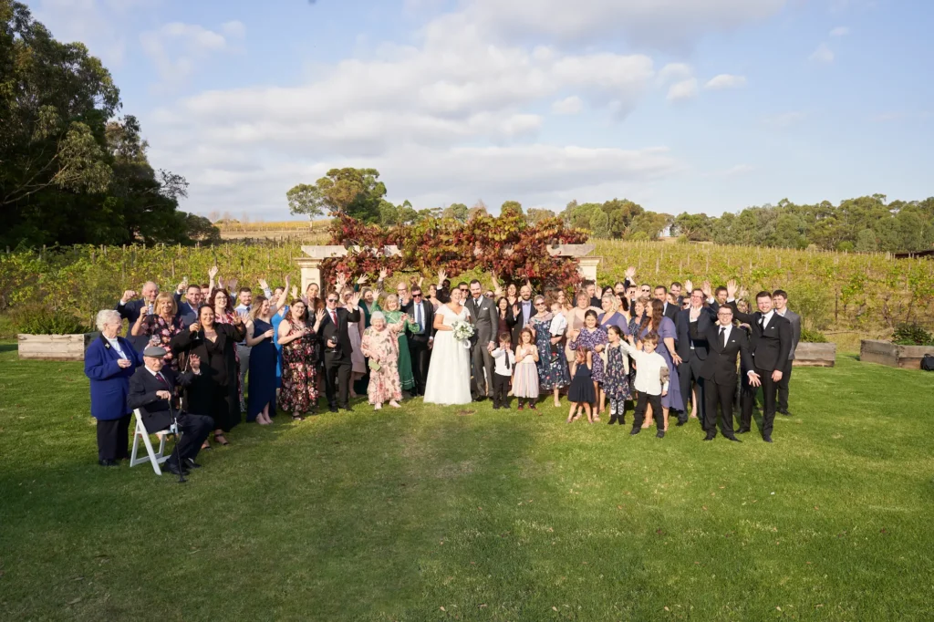 erin jesse family group photo fergusson winery yarra valley wedding A joyful wedding group photo at Fergusson Winery, with bride and groom Erin and Jesse at the center surrounded by family and friends. Guests of all ages are arranged in multiple rows on a lush green lawn, some waving enthusiastically, others smiling warmly at the camera. Behind them, the Sandstone Gazebo is draped in vibrant red and green grapevines, with vineyards and rolling hills stretching into the distance. The bright blue sky and natural light create a cheerful, celebratory, and heartfelt atmosphere, capturing the collective happiness of everyone who attended the wedding.