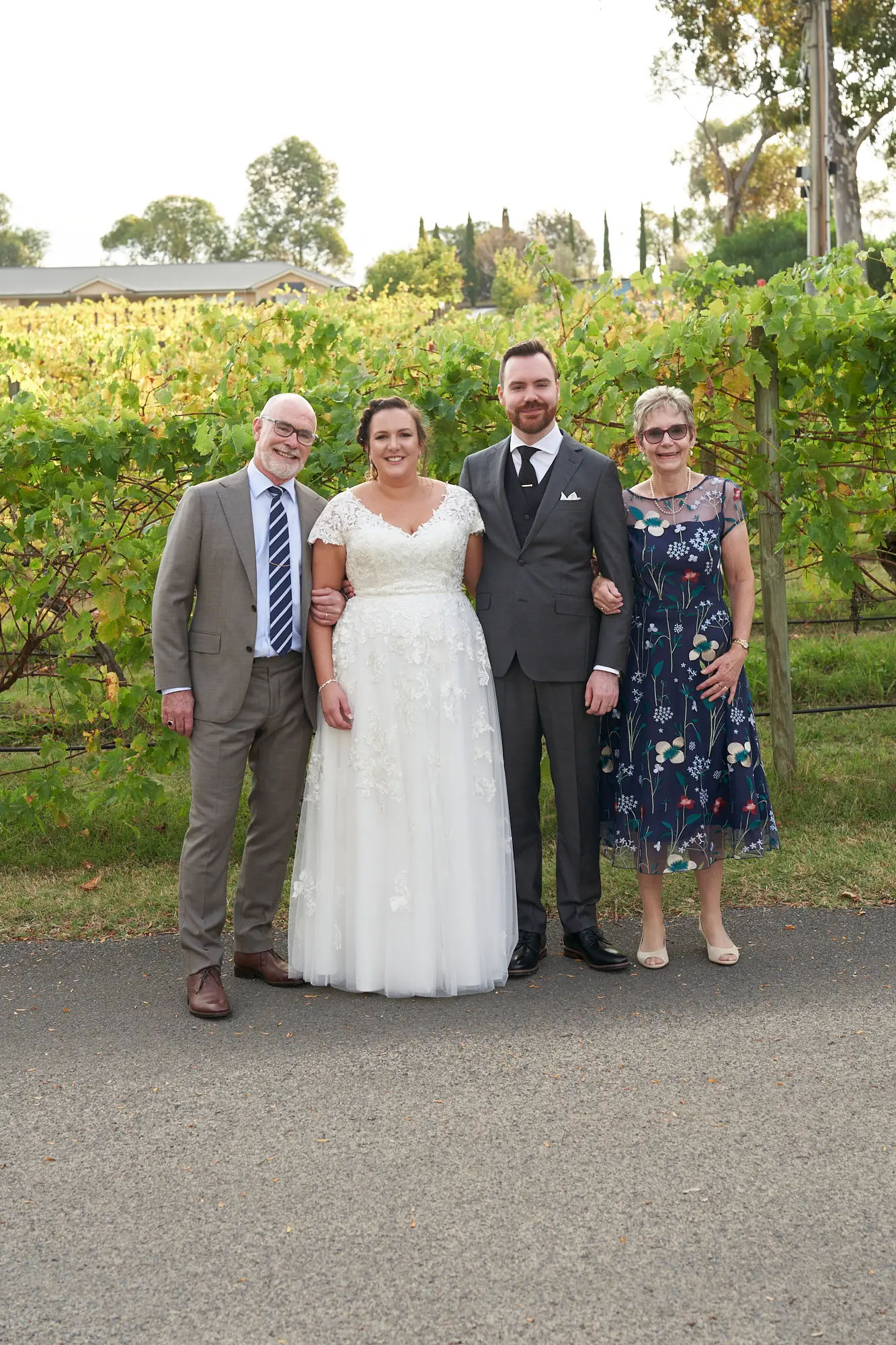 erin jesse family portrait groom parents fergusson winery yarra valley wedding Bride and groom Erin and Jesse pose outdoors with the groom's parents at Fergusson Winery. Standing side-by-side on a paved path, the couple is centered, smiling warmly, with the groom’s arm around the bride’s waist and the bride linked with his father. The parents smile proudly beside them. Behind them, rows of grapevines in vibrant green and yellow autumn tones stretch into the distance, with rolling hills of the Yarra Valley visible. The natural light and vineyard setting create a joyful, elegant, and celebratory family portrait capturing this special wedding day.