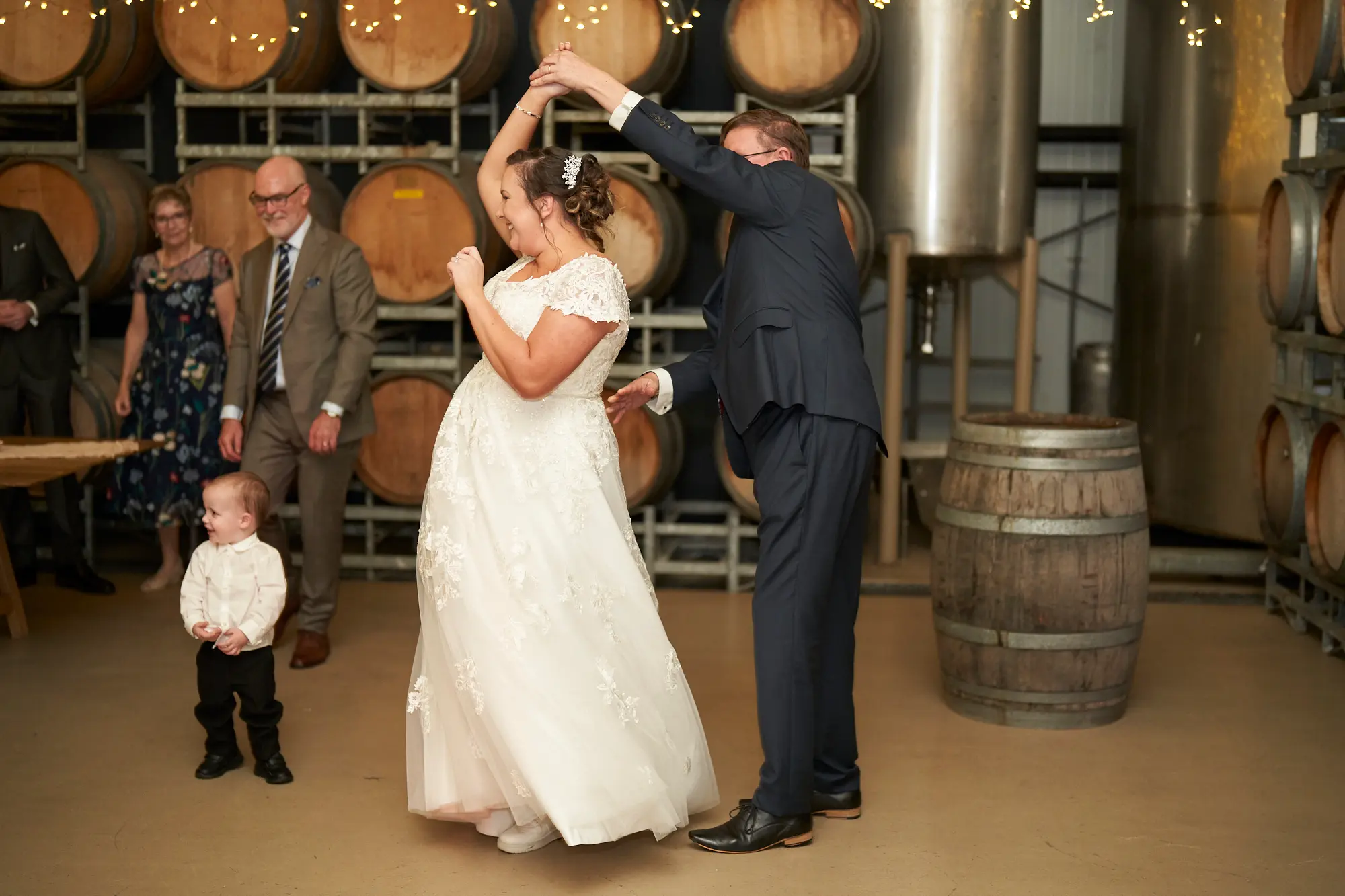 erin jesse father daughter dance barrel room fergusson winery yarra valley wedding The bride joyfully dances with her father in Fergusson Winery’s barrel room, her hands lifted as he spins her with a proud smile. A young boy watches from the foreground while seated guests smile in the background. Warm string lights glow above the polished concrete floor, illuminating rows of stacked French oak barrels and a stainless steel wine tank. The moment feels lively, intimate, and full of heartfelt celebration, capturing the happiness of their father-daughter dance within the rustic-chic charm of the Yarra Valley winery setting.