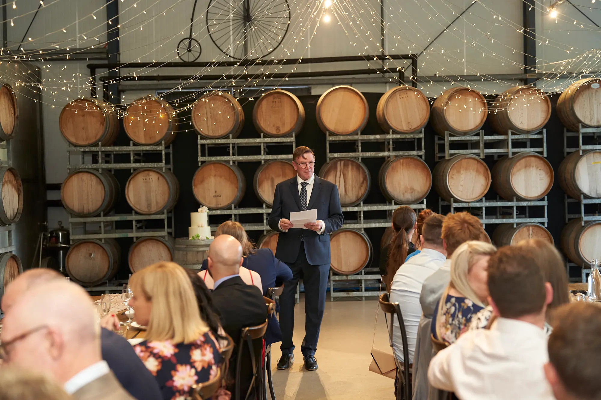 erin jesse father of bride speech barrel room fergusson winery A touching moment at Fergusson Winery’s barrel room as the bride’s father gives a heartfelt speech during the wedding reception. Guests sit at long, rustic wooden tables, listening attentively, bathed in the warm glow of hundreds of twinkling string lights overhead. Stacked oak wine barrels line the walls, adding rustic charm, while a two-tiered wedding cake rests nearby, ready for later celebration. The scene captures an intimate, emotional, and elegant atmosphere, highlighting the connection between the speaker and the guests and the cozy, inviting ambiance of the winery’s barrel room.