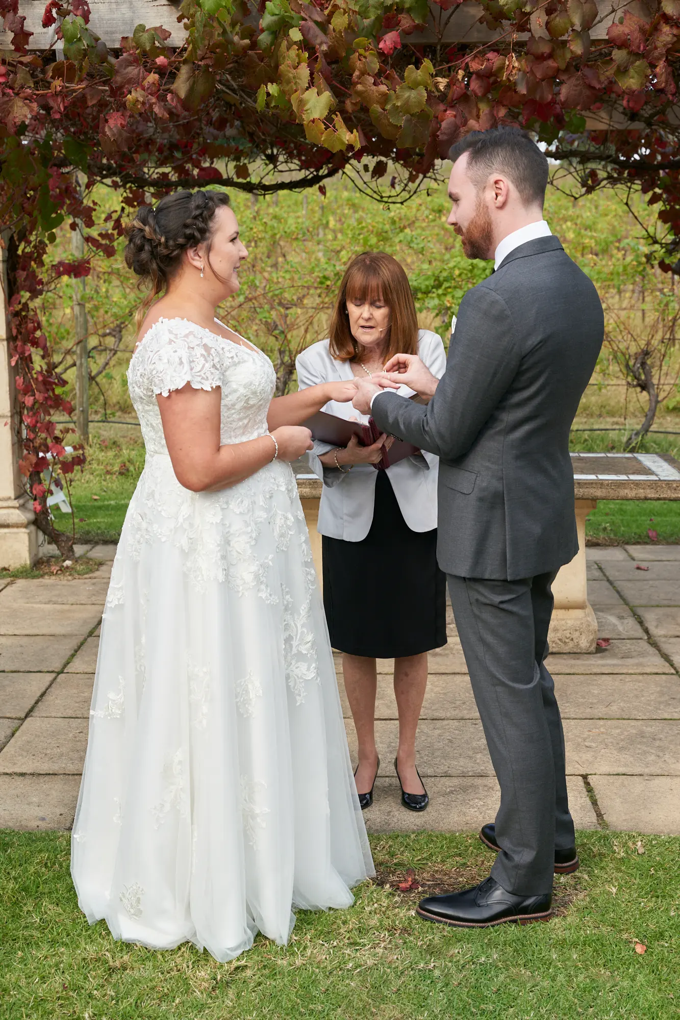 erin jesse groom placing ring ceremony fergusson winery yarra valley Groom placing a wedding ring on the bride’s left finger beneath a rustic wooden pergola draped in vibrant red and green autumn grapevines at Fergusson Winery in the Yarra Valley. The bride looks toward him with a gentle smile as the officiant stands between them holding a book to guide the ceremony. Rows of vineyards stretch into the distance behind them, with stone columns, paved ground, and green lawn completing the serene, rustic, and elegant outdoor setting. This intimate moment captures the joy, focus, and emotional significance of their ring exchange.