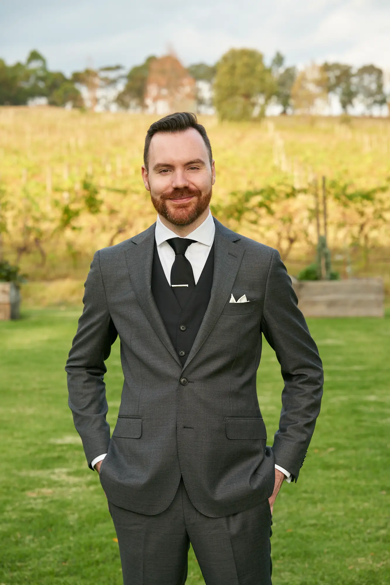 erin jesse groom portrait fergusson winery yarra valley wedding A confident groom poses outdoors at Fergusson Winery in the Yarra Valley, standing on a lush green lawn with hands casually in his suit pockets. Dressed in a dark gray suit with a black waistcoat, white shirt, black tie, and pocket square, he smiles warmly at the camera. Behind him, rows of grapevines and rolling hills create a scenic vineyard backdrop, blending rustic natural beauty with the groom’s formal elegance and capturing the joyful, celebratory atmosphere of the wedding day.