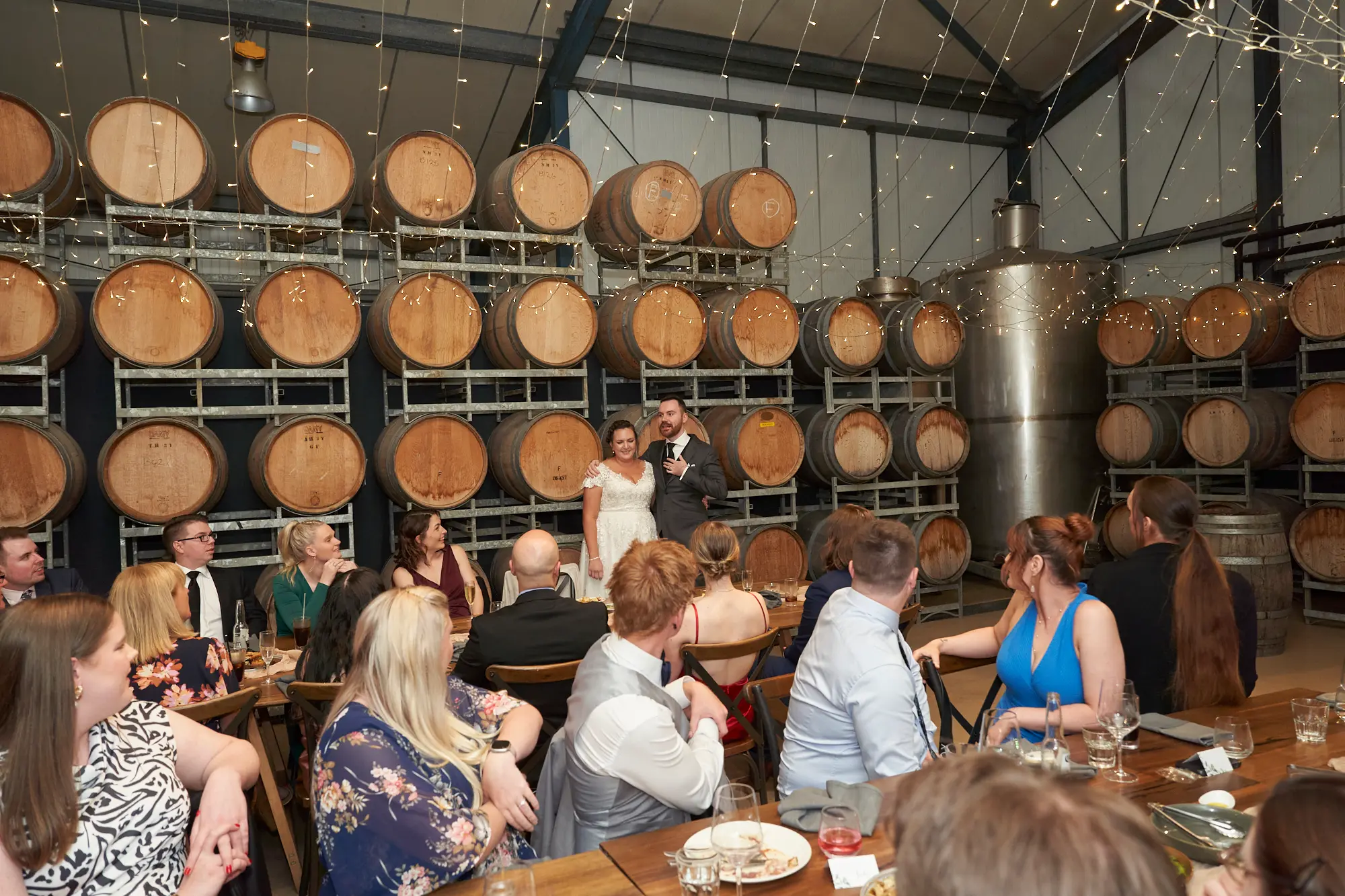 erin jesse groom speech barrel room fergusson winery yarra valley wedding The groom delivers a warm and joyful speech during the wedding reception at Fergusson Winery’s barrel room, with the bride standing closely beside him, smiling. Guests seated at rustic wooden tables listen attentively as stacked wine barrels and a large stainless steel tank frame the room. Hundreds of twinkling string lights overhead cast a soft, intimate glow, creating a cozy, celebratory, and rustic-chic atmosphere that highlights the personal and heartfelt moment of the winery wedding reception.