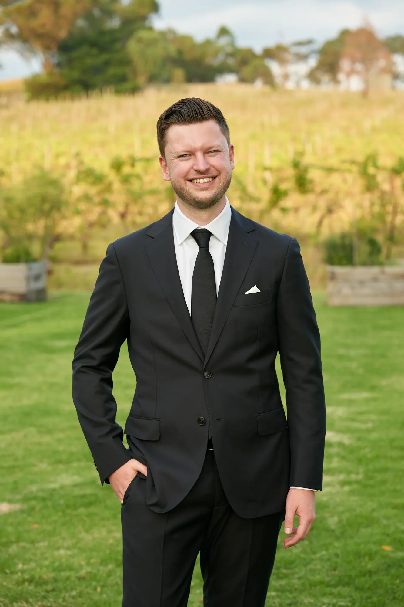 erin jesse groomsman portrait fergusson winery yarra valley wedding A confident groomsman poses outdoors at Fergusson Winery in the Yarra Valley, standing on a lush green lawn with his right hand casually in his suit pocket. He wears a black suit, white shirt, black tie, and pocket square, smiling warmly at the camera. Behind him, rows of grapevines and rolling hills form a scenic vineyard backdrop, combining rustic natural beauty with formal elegance, capturing the celebratory and joyful atmosphere of the wedding day.