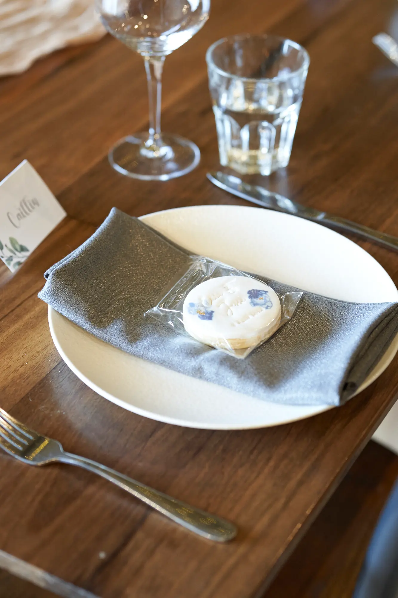 erin jesse guest table setting fergusson winery This photo captures an intimate and elegant guest table setting at Fergusson Winery’s barrel room. A dark wooden table is set with a white plate, gray cloth napkin, silver cutlery, and two clear glasses. A personalized sugar cookie and handwritten name card reading “Caillin,” adorned with green and blue floral designs, add a thoughtful, welcoming touch. Soft lighting highlights the warm, rustic textures of the wood, creating a cozy, personalized atmosphere. The combination of refined dinnerware with the natural, earthy elements of the winery venue exudes charming rustic elegance, ready for guests to enjoy the wedding reception.