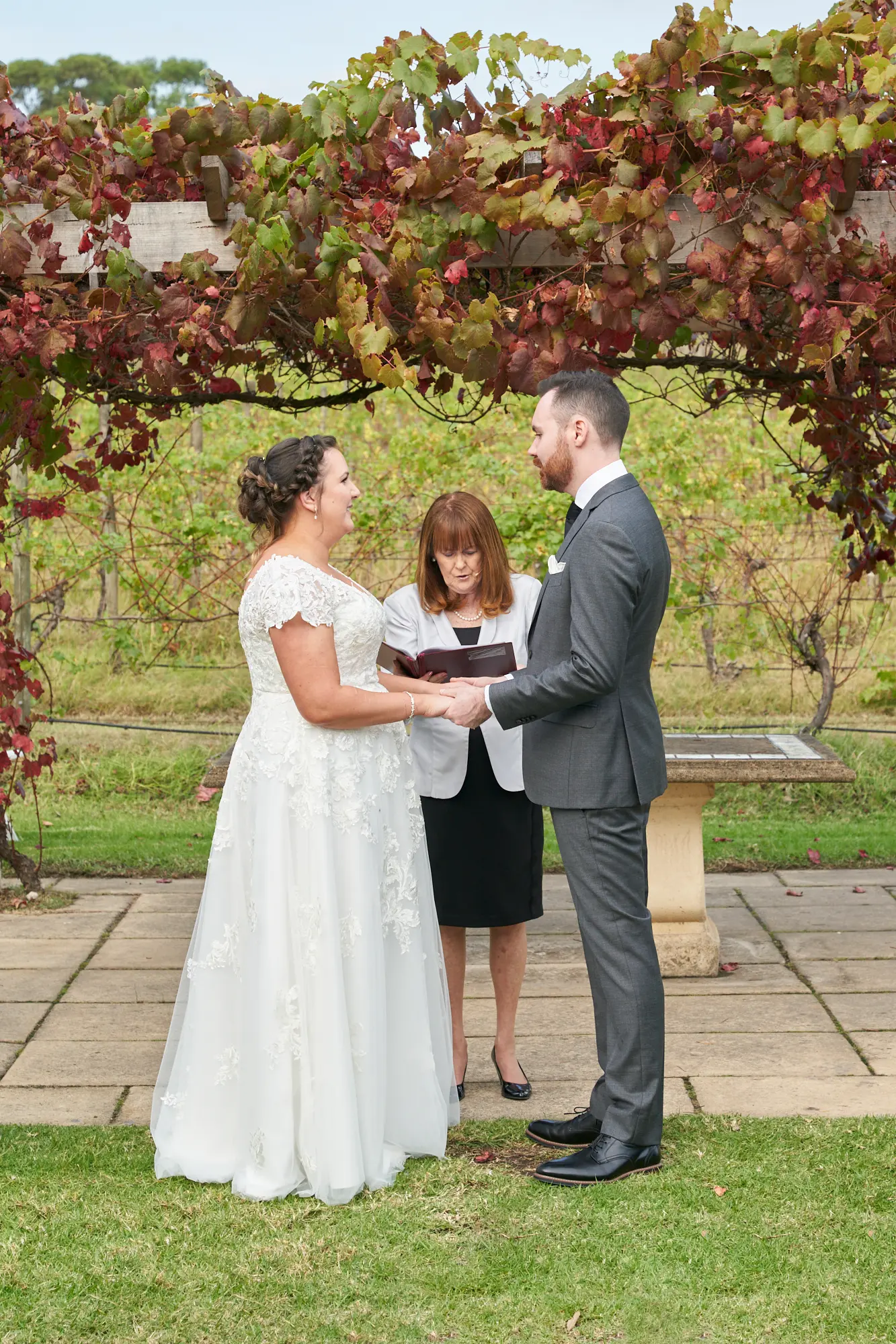 erin jesse holding hands fergusson winery yarra valley Bride and groom holding hands beneath a wooden pergola draped in vibrant red and green autumn grapevines during their outdoor wedding ceremony at Fergusson Winery in the Yarra Valley. They face each other with focused, heartfelt expressions as the officiant stands between them, guiding the vow exchange. Rows of vineyards stretch into the background under a bright sky with scattered clouds, creating a serene and rustic setting. The moment feels intimate, solemn, and joyfully elegant, blending natural beauty with the emotional significance of the ceremony.