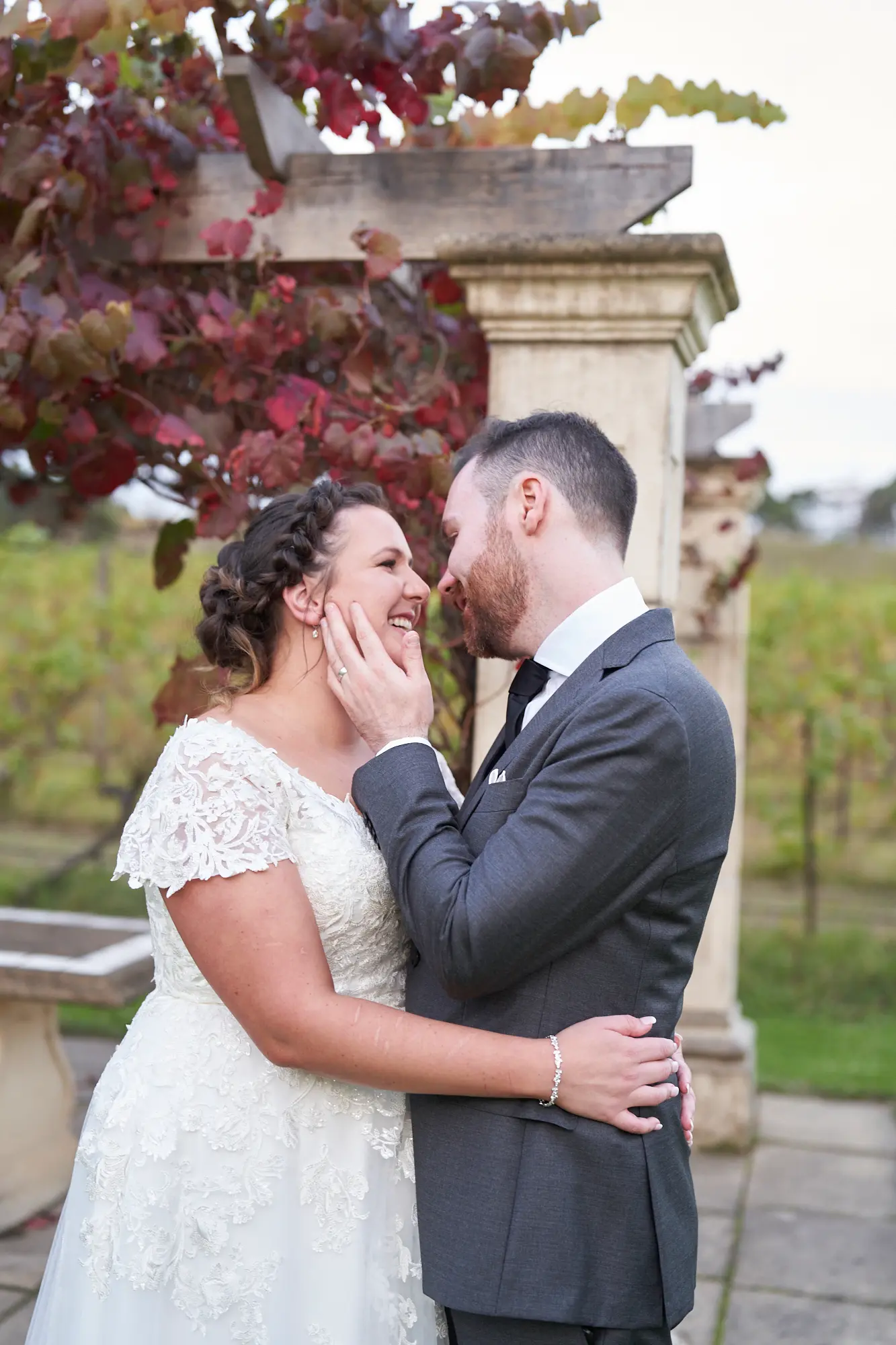 erin jesse intimate gaze sandstone gazebo fergusson winery yarra valley wedding This wedding photo captures a romantic and intimate moment between the bride and groom at Fergusson Winery in the Yarra Valley. Standing close under the vine-draped sandstone gazebo, they gaze into each other’s eyes with joyful smiles, the groom’s arm around the bride’s waist and her hands on his cheeks. Autumn-toned grapevines frame them naturally, while vineyards and rolling hills stretch in the background. The serene, heartfelt portrait conveys deep emotional connection, warmth, and rustic elegance, capturing a private, unguarded moment of love and happiness in a peaceful outdoor setting.